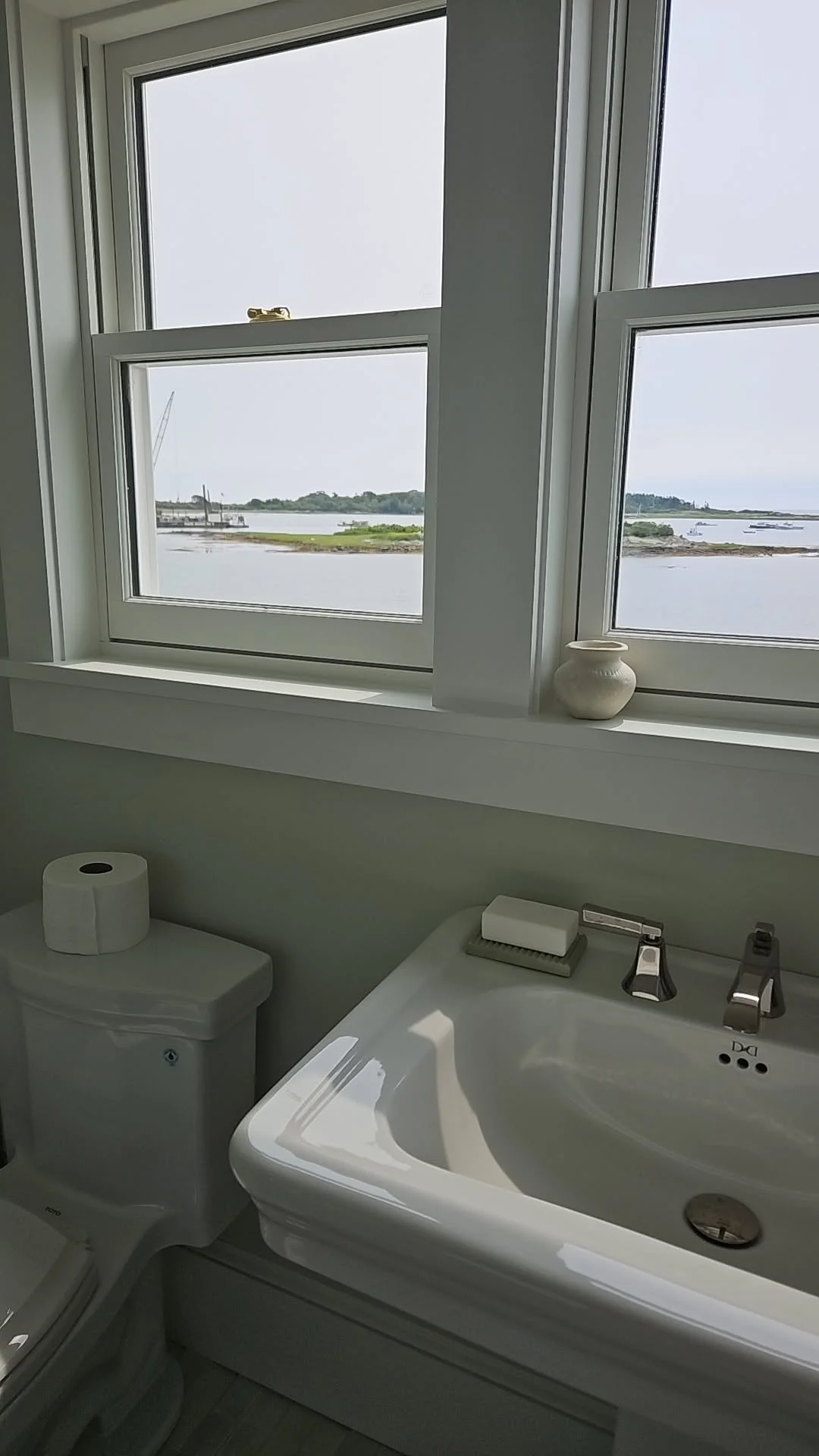 Bathroom with large window overlooking a body of water and distant shoreline, white bathtub, toilet with toilet paper roll, soap on edge of tub, small white vase on window sill.