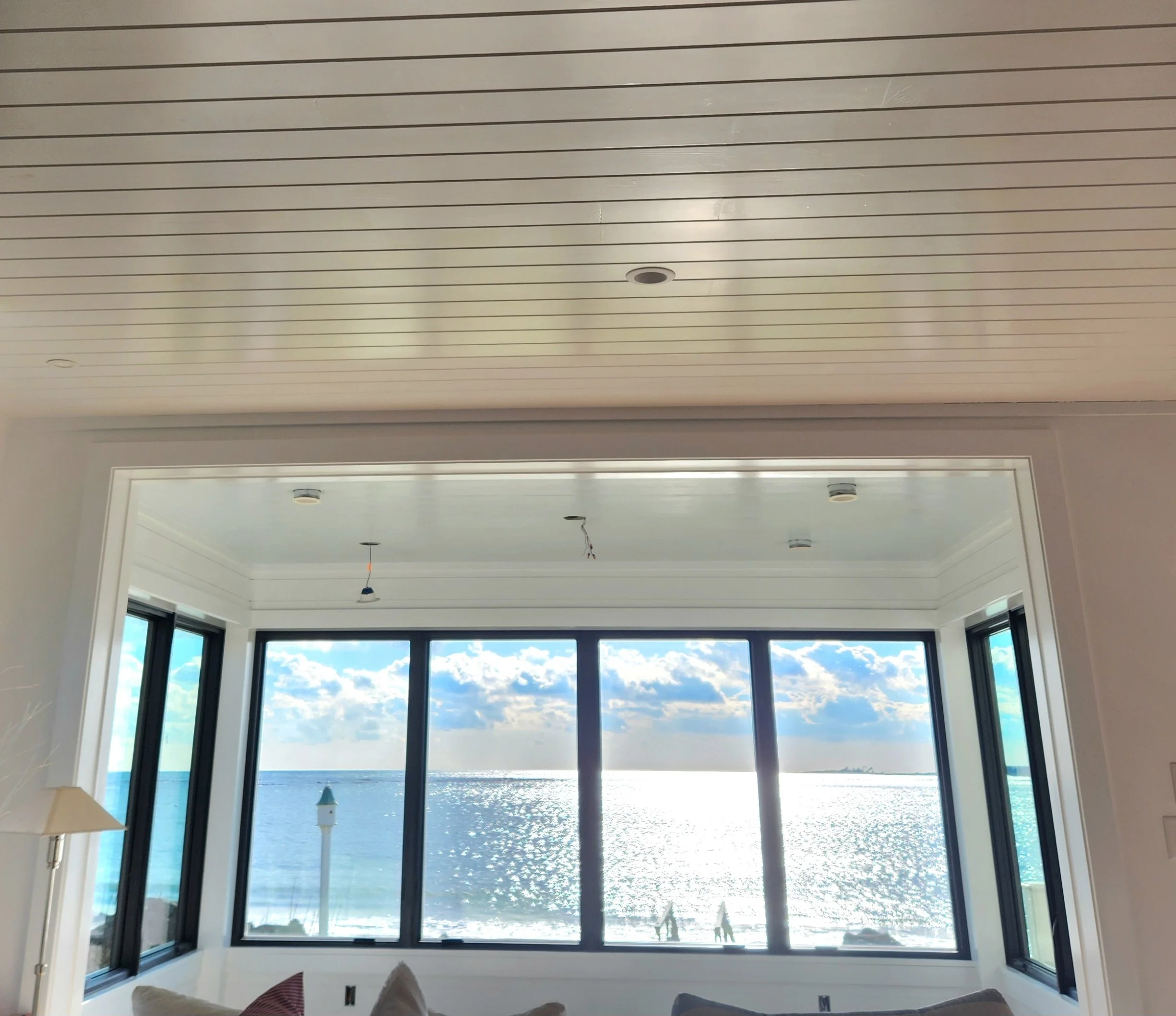 Living room with large window overlooking a body of water and sky with clouds, featuring a ceiling with parallel lines and a partial view of a beige sofa and a table lamp.