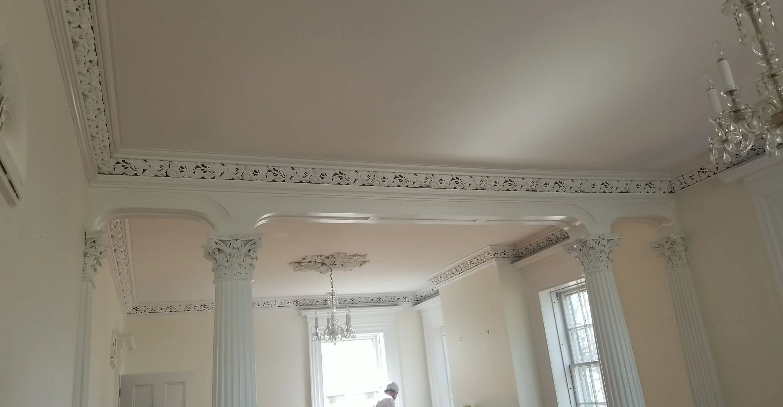 Interior of a room with white ornate crown molding, decorative columns, a chandelier, and large windows.