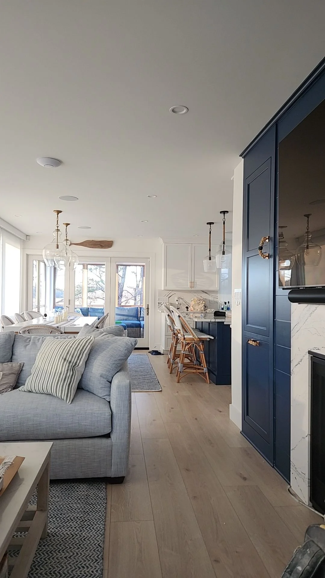 Open-concept living room and kitchen with light wood flooring, light-colored sofa with striped pillows, blue kitchen cabinets, bar seating with wicker stools, and large windows and glass doors letting in natural light.