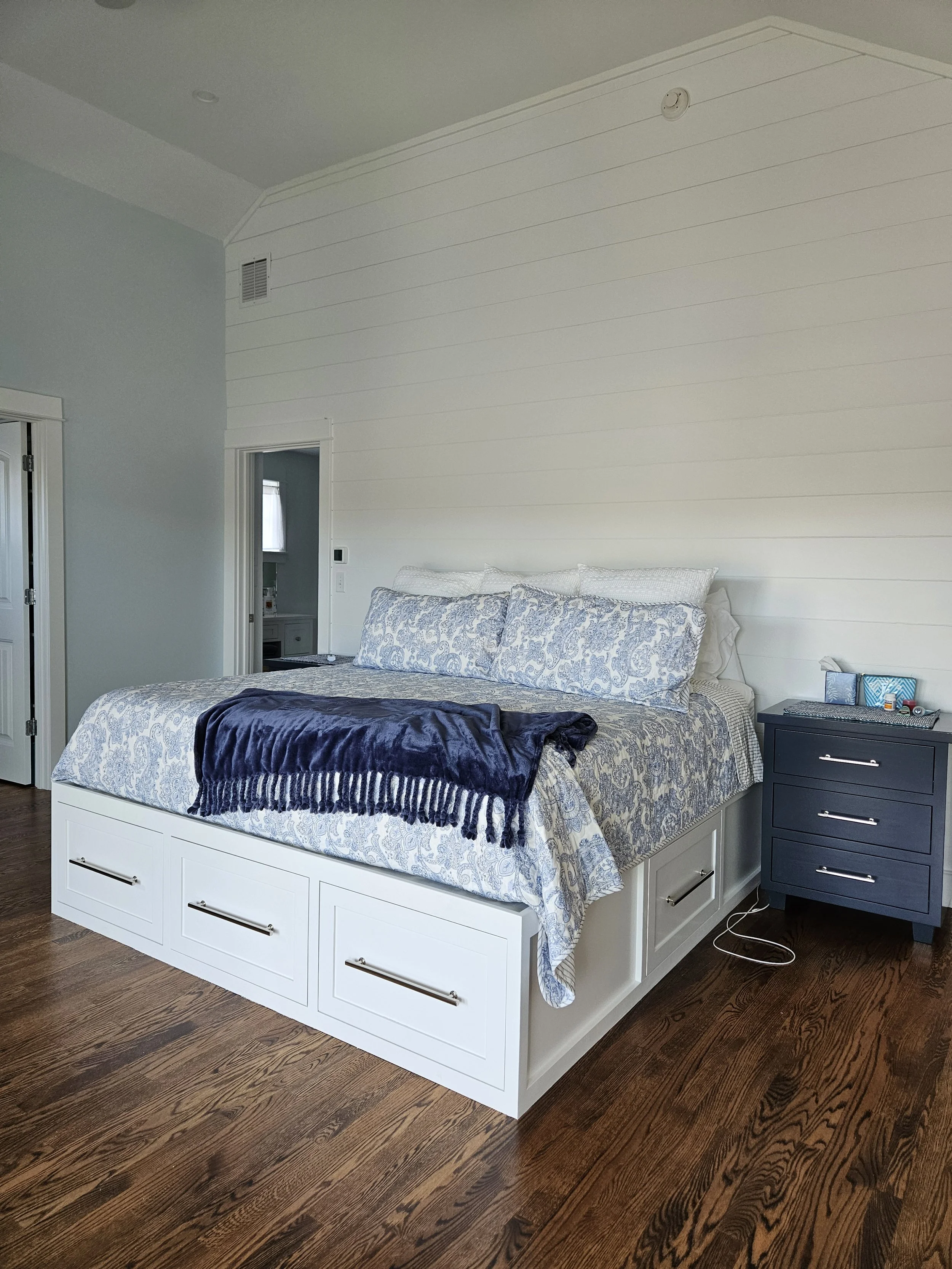 A bedroom with a white bed frame, patterned bedding, and a blue nightstand. The bed has multiple pillows, and a dark blanket is draped over part of it. The room has wooden floors, and a door to a closet is visible on the left. There is a push-button 