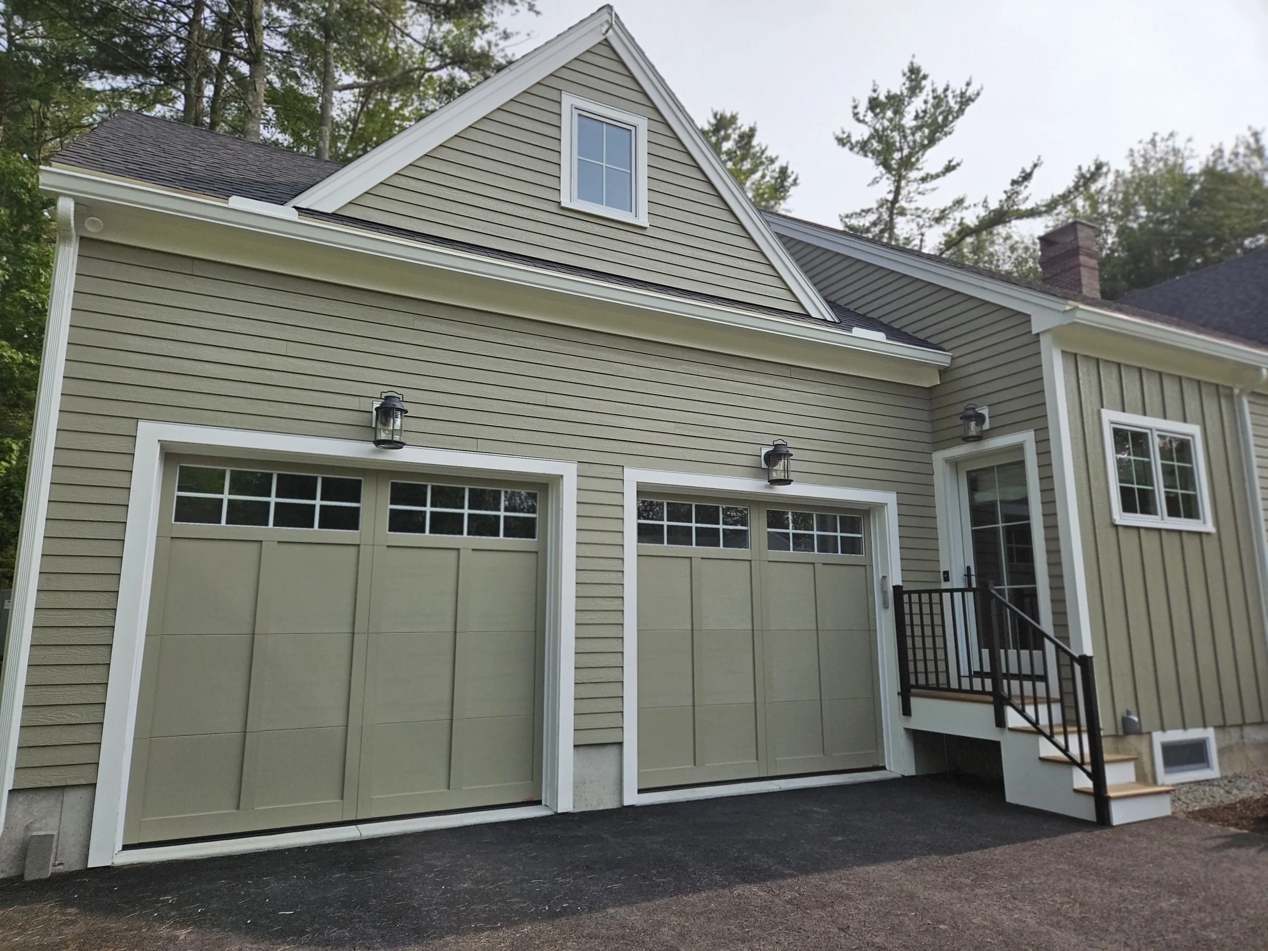 The image shows the exterior of a house with beige siding and white trim. There is a large garage door at the bottom, a window on the second floor, exterior lighting fixtures, and a staircase with wooden steps and black railing leading to a door.