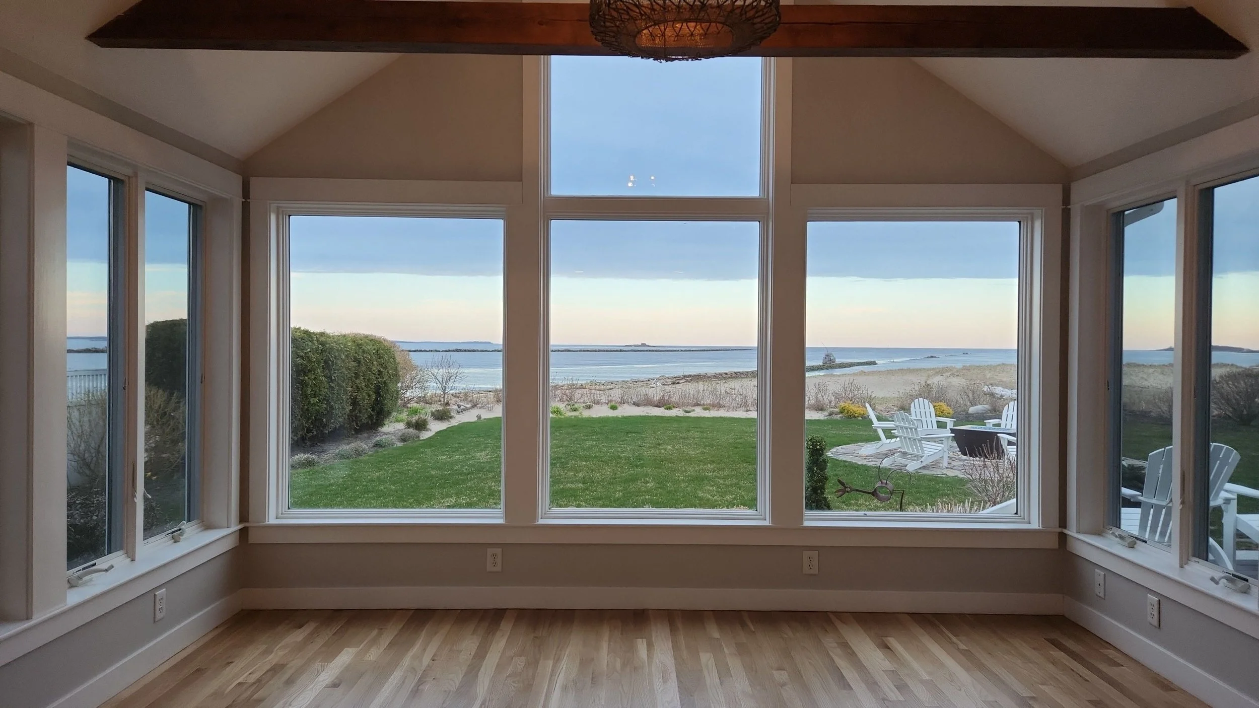 Room with large window view of a beach, grassy yard, and patio with white chairs and landscaping.