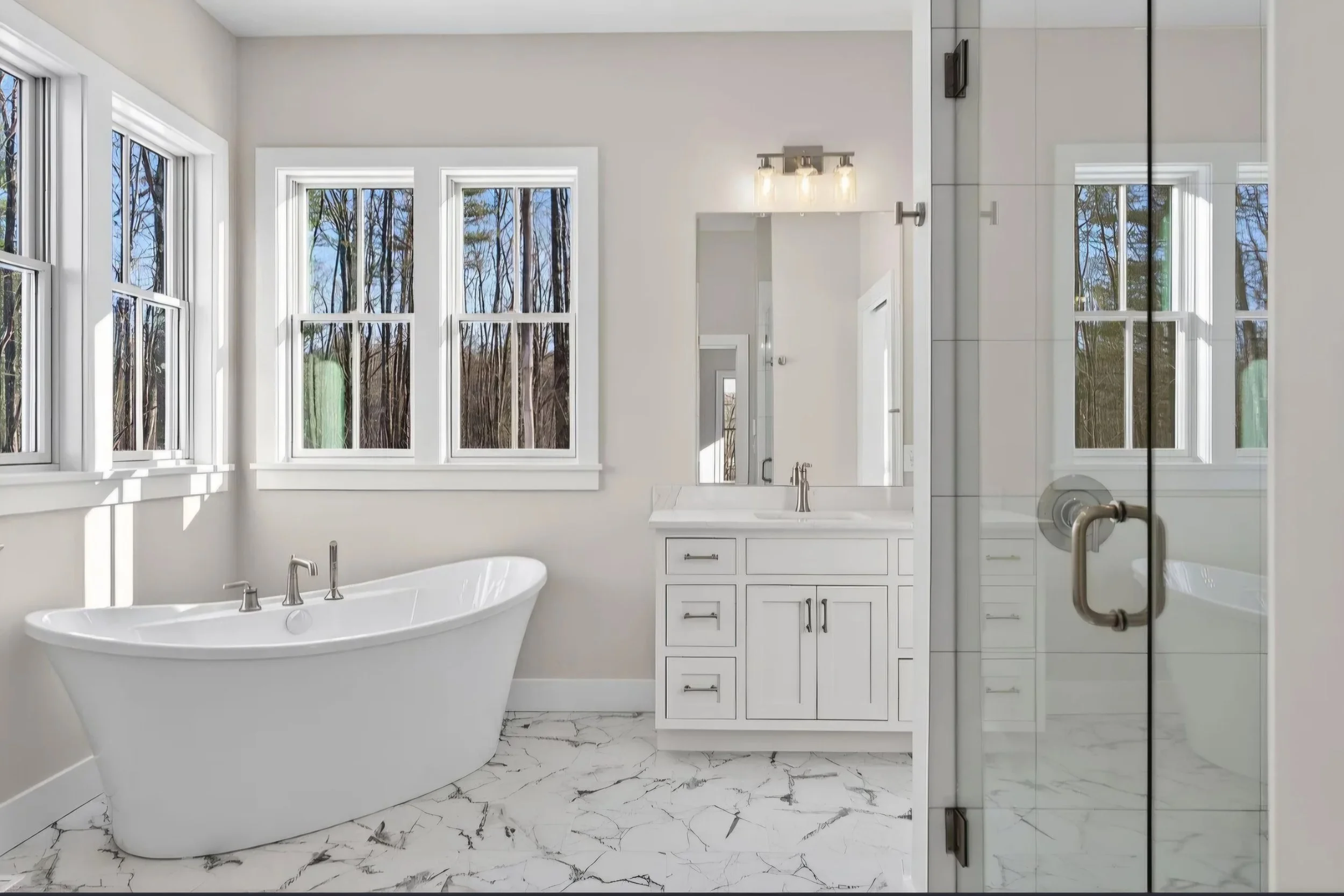 Bright white bathroom with large windows showing trees outside, a freestanding bathtub, white cabinetry, and marble-patterned floor tiles.