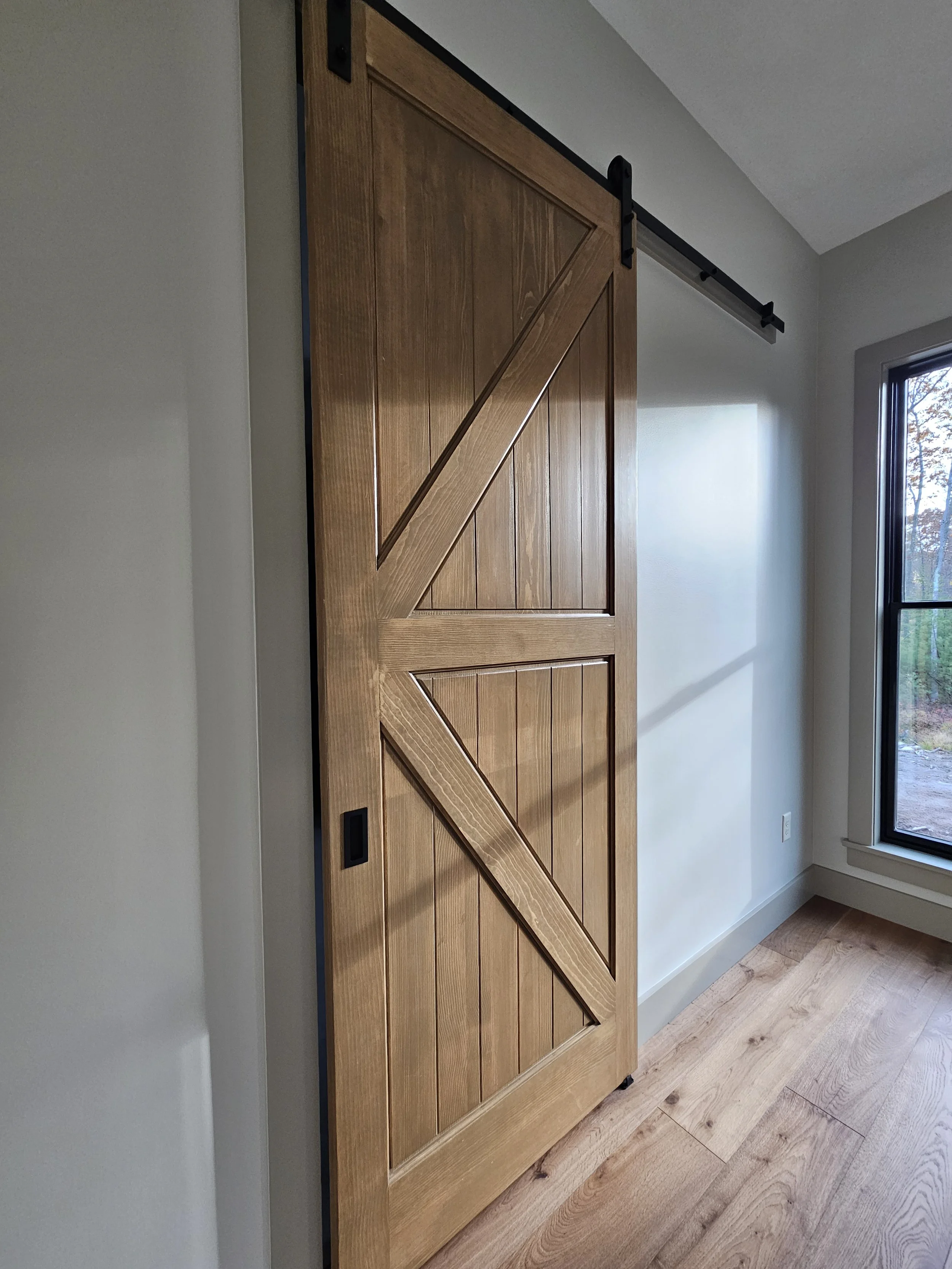 A wooden sliding barn door on a black metal rail installed at the top of the doorway, with a natural wood finish, in a room with light-colored walls and a window with a view of trees outside.