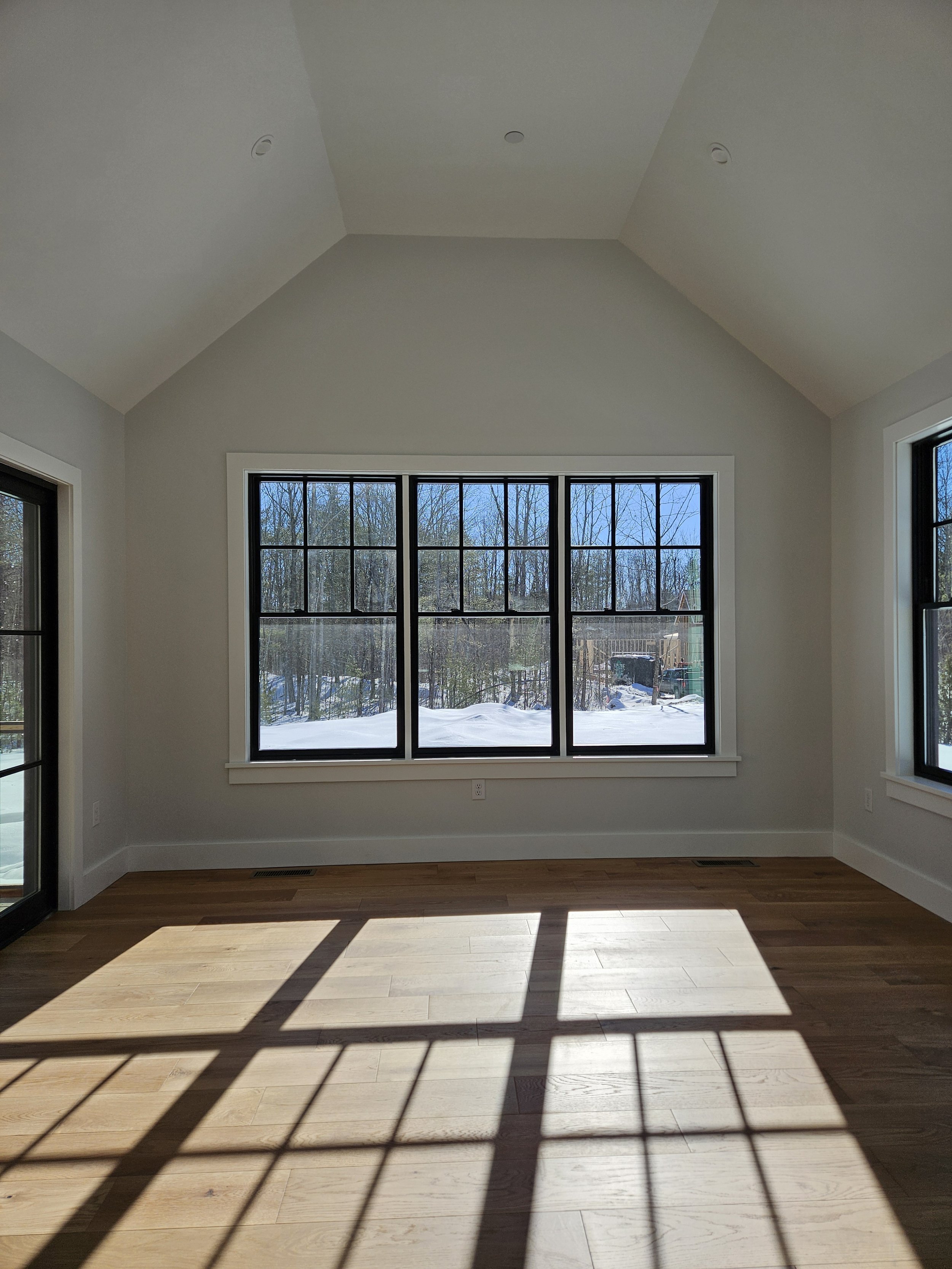 Empty room with large windows showing snowy outdoors, sunlight casting shadows on wooden floor.