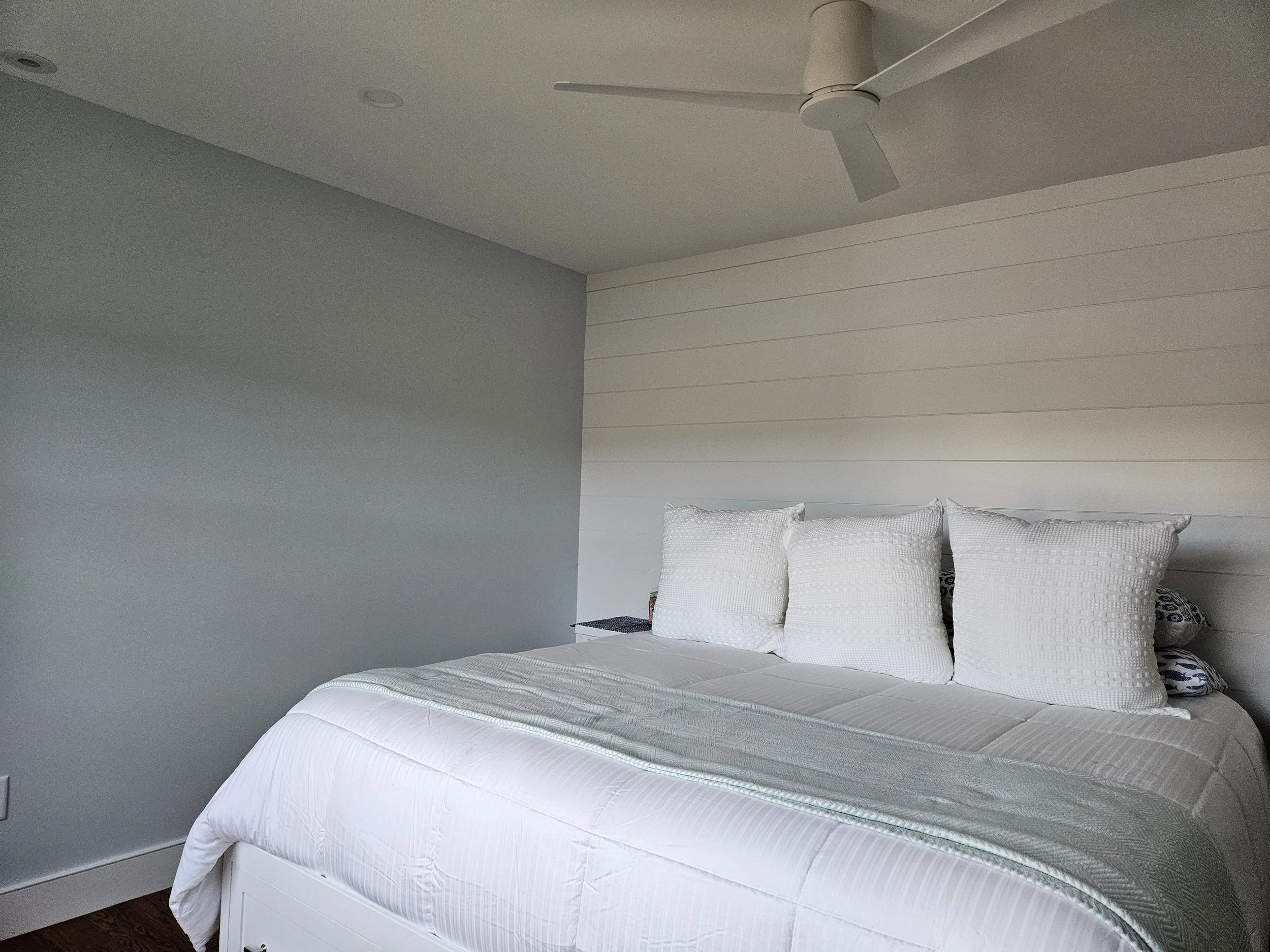 A bedroom with a white bed, pillows, wall-mounted shelves, a white wall, and a ceiling fan.