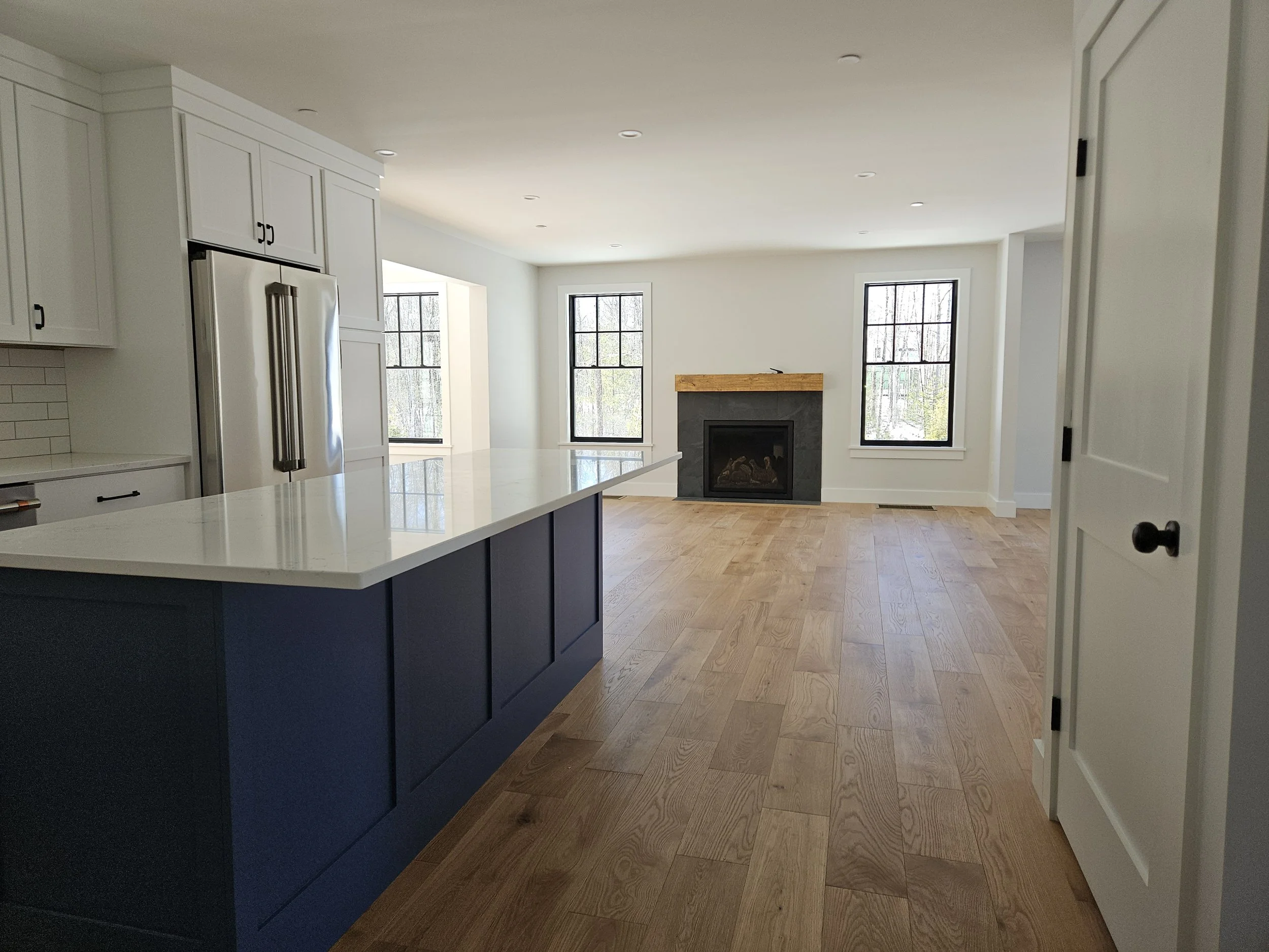 Empty bright living room with three large windows, white walls, wood flooring, a fireplace with a wooden mantle, and a kitchen island with dark blue cabinetry.