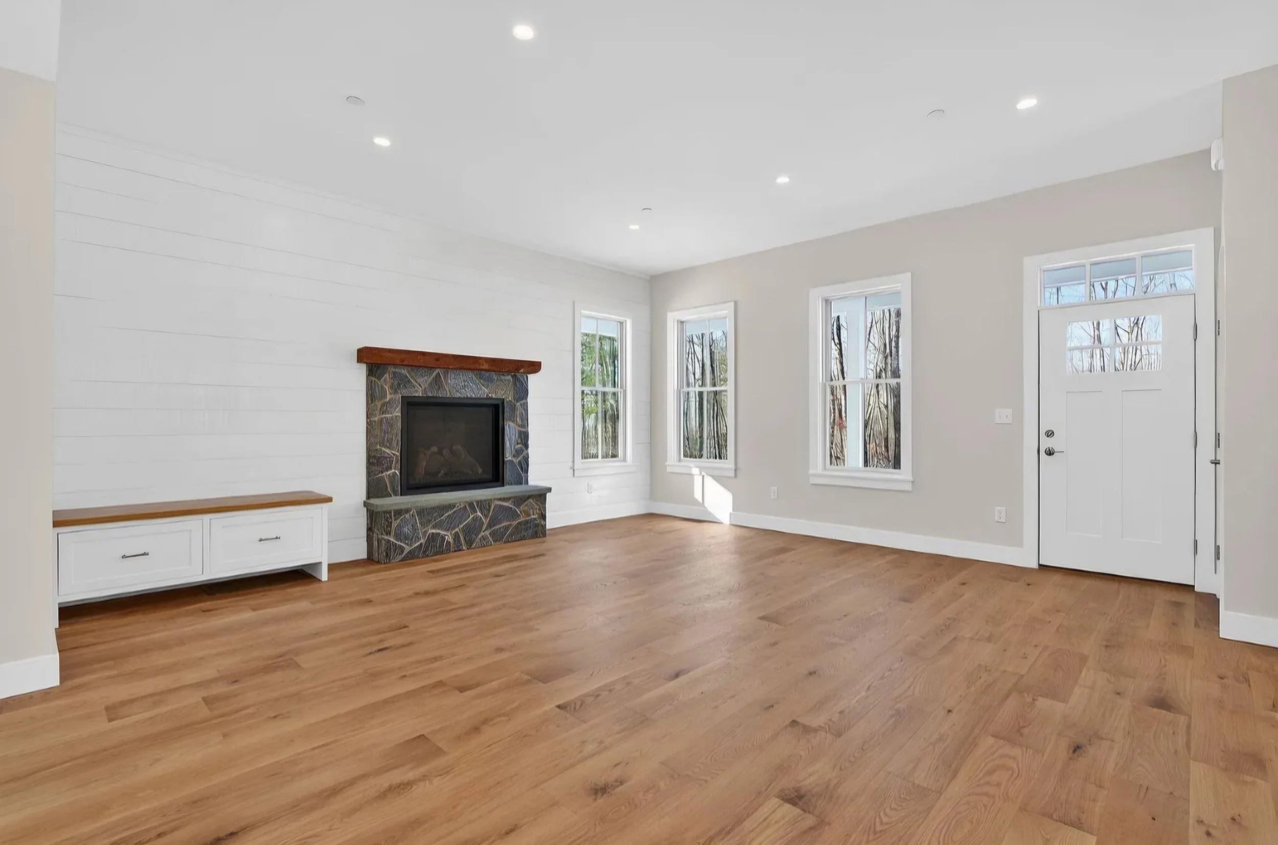 Empty living room with hardwood floors, white walls, three windows, a fireplace with a wooden mantel, white cabinet, and a door, illuminated by ceiling lights.