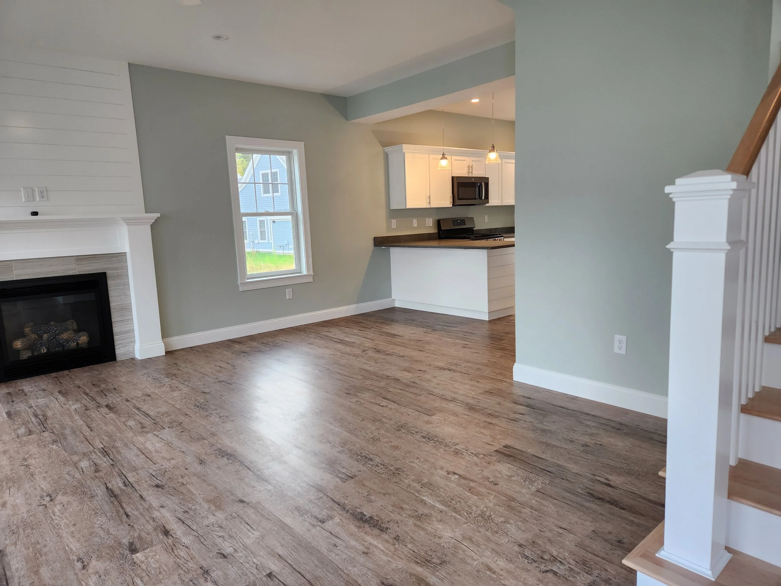 Open-concept living room with fireplace, hardwood flooring, light gray walls, and a view into the kitchen with white cabinetry and pendant lighting.