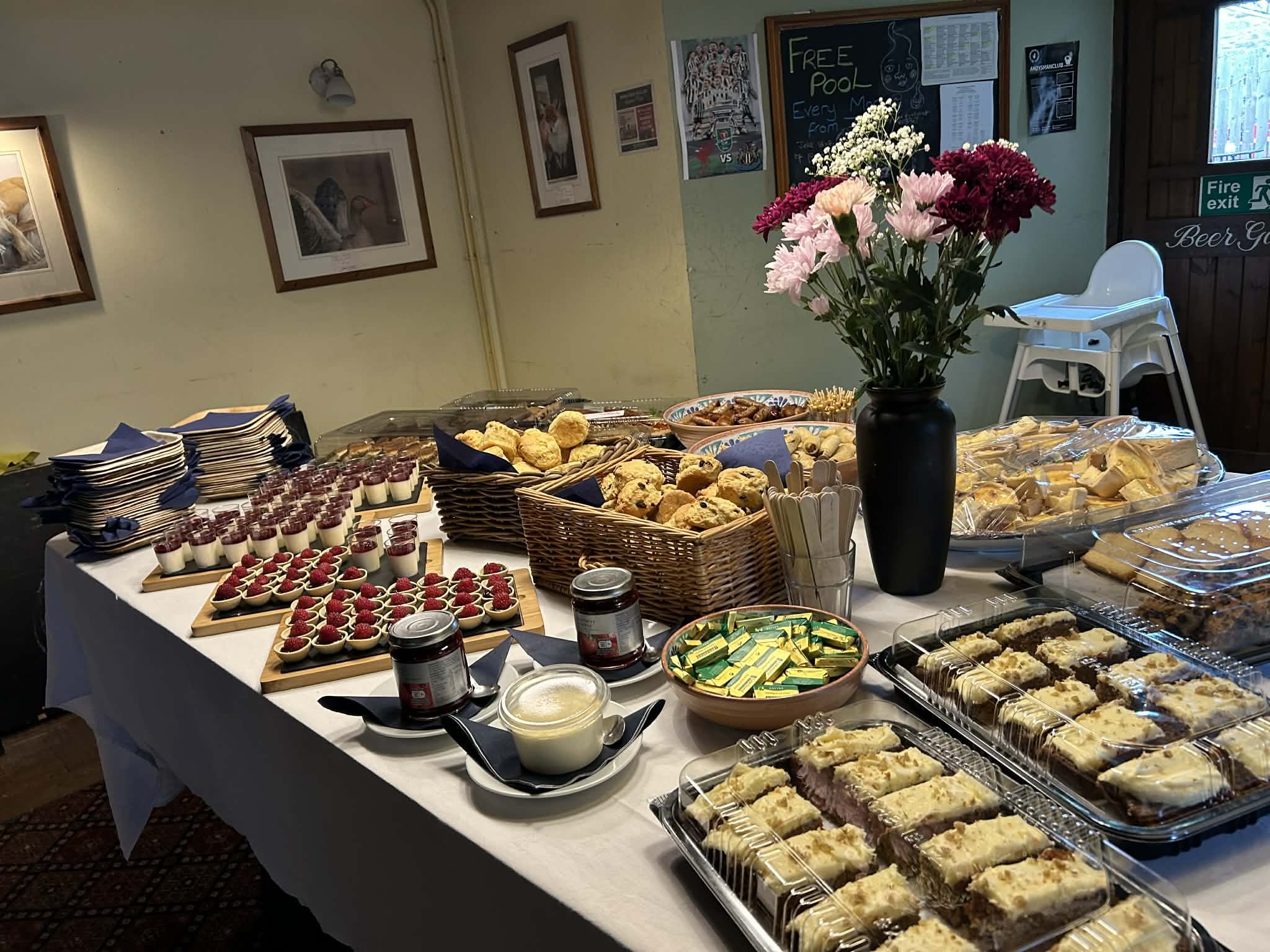 A buffet table with various desserts such as parfaits topped with raspberries, cookies, brownies, and other baked goods. In the background, a black vase with fresh pink and burgundy flowers, posters on the wall, and a chalkboard advertising a free pool are visible.