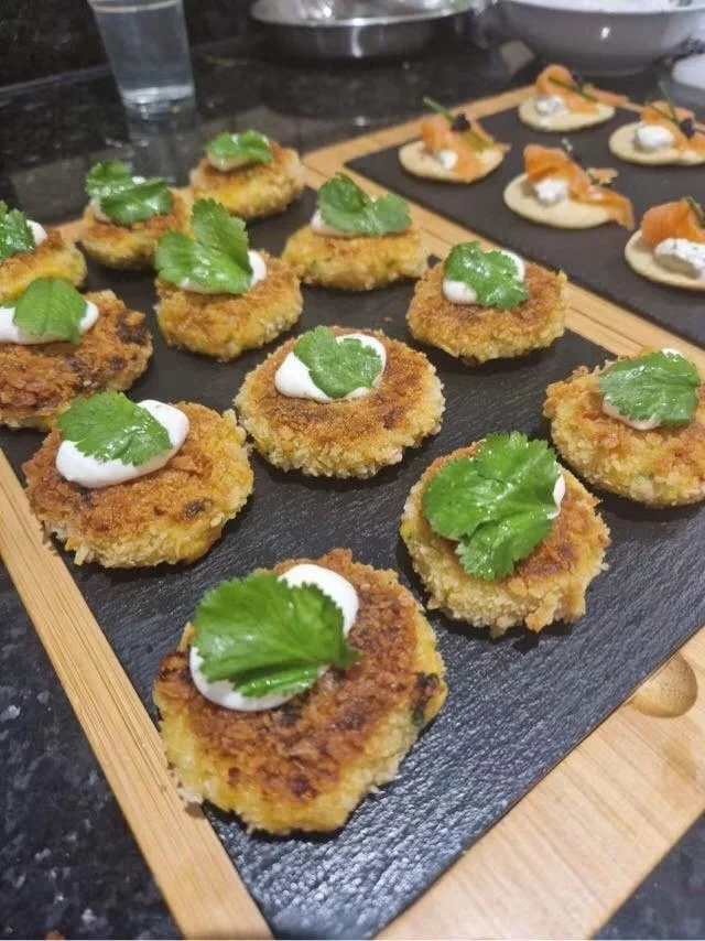 Close-up of breaded, fried appetizers topped with green herbs and creamy sauce, arranged on a black serving board.