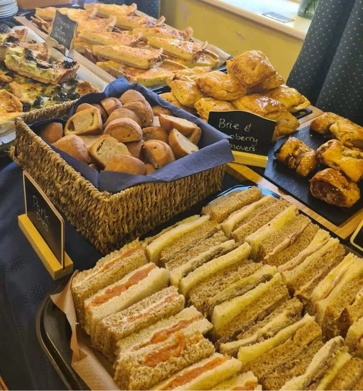 Assorted baked goods including finger sandwiches, scones, pastries, and fruit-filled danishes displayed on trays in a cafe or bakery setting.