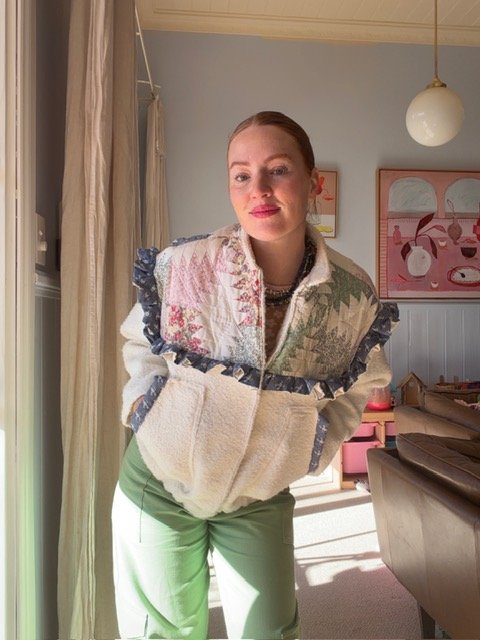 A woman standing inside a room, leaning slightly forward, with light coming in from the left. She wears a colorful patterned jacket with ruffled sleeves over a cream sweater, and green pants. The room has pastel-colored artwork on the wall and a pend
