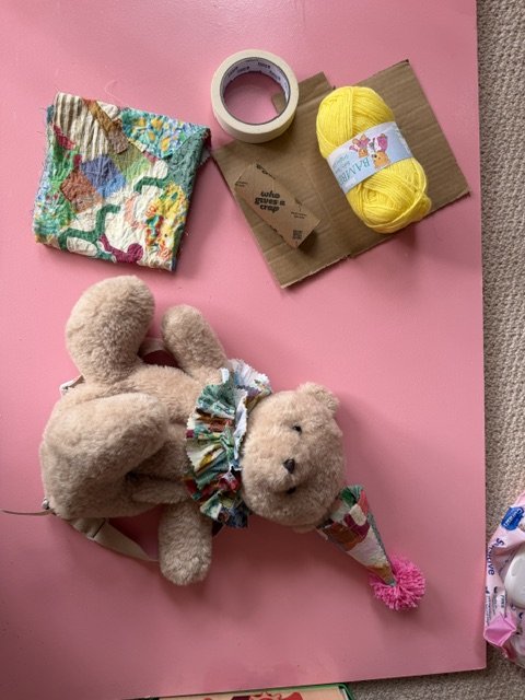 A pink table with craft supplies and a stuffed teddy bear wearing a colorful ruffled collar and party hat. The supplies include a multicolored knitted cloth, a roll of tape, a ball of yellow yarn, a piece of cardboard, and a small piece of brown pape