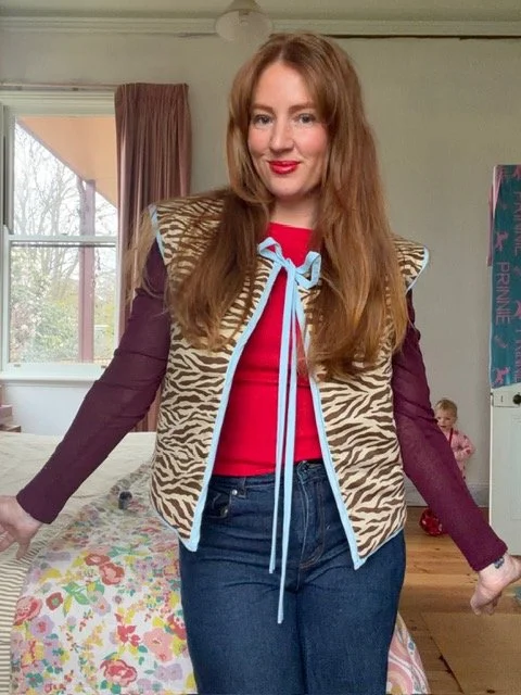 A woman with long red hair wearing a patterned vest with zebra stripes and shoulder pads, a red top with a blue ribbon, and blue jeans standing in a bedroom.