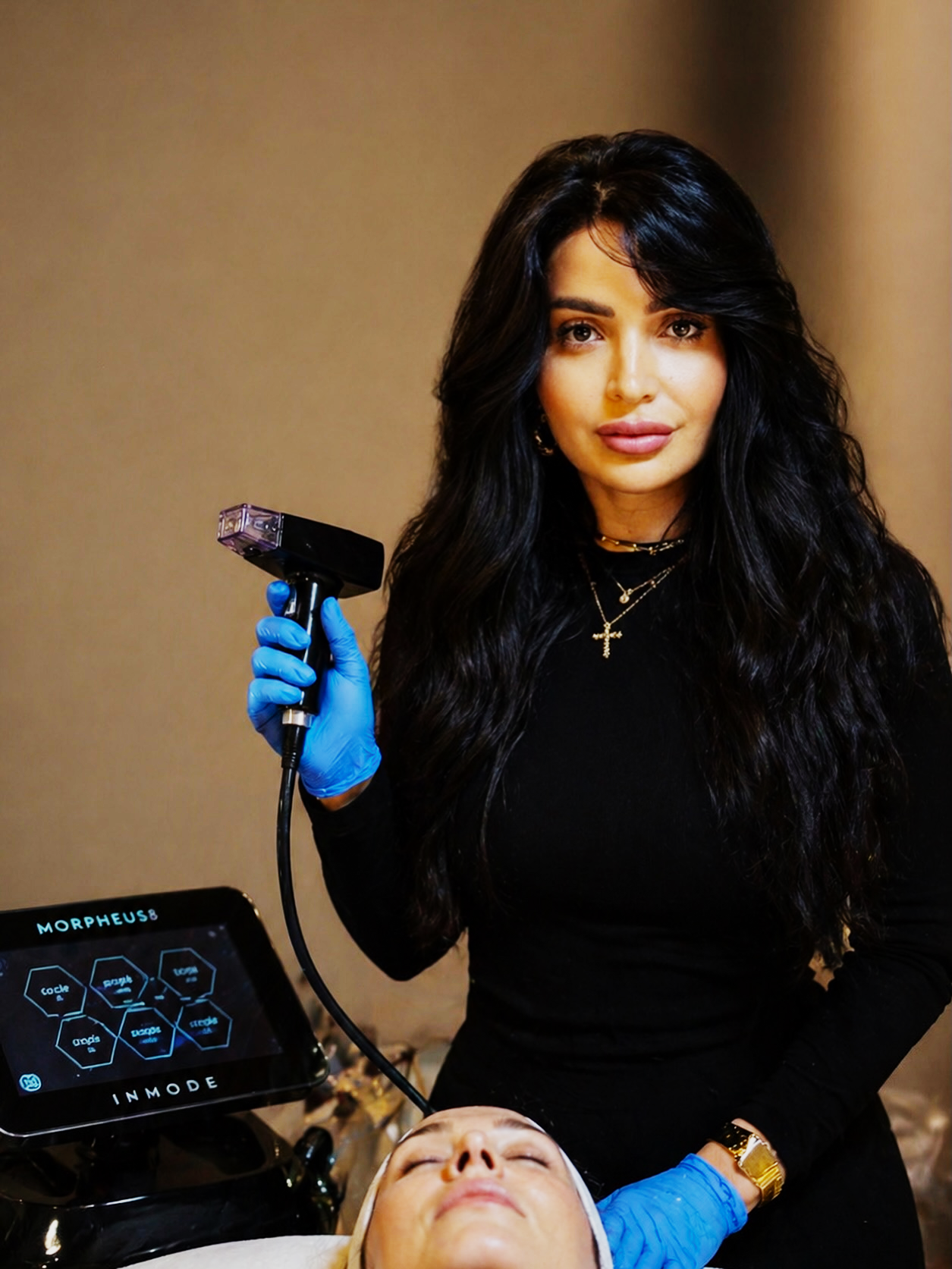 A woman with long black hair, wearing a black shirt, gold jewelry, and blue gloves, holding a laser tattoo removal device near a person's face, who is lying down with eyes closed. Medical equipment with a digital display is visible in the foreground.