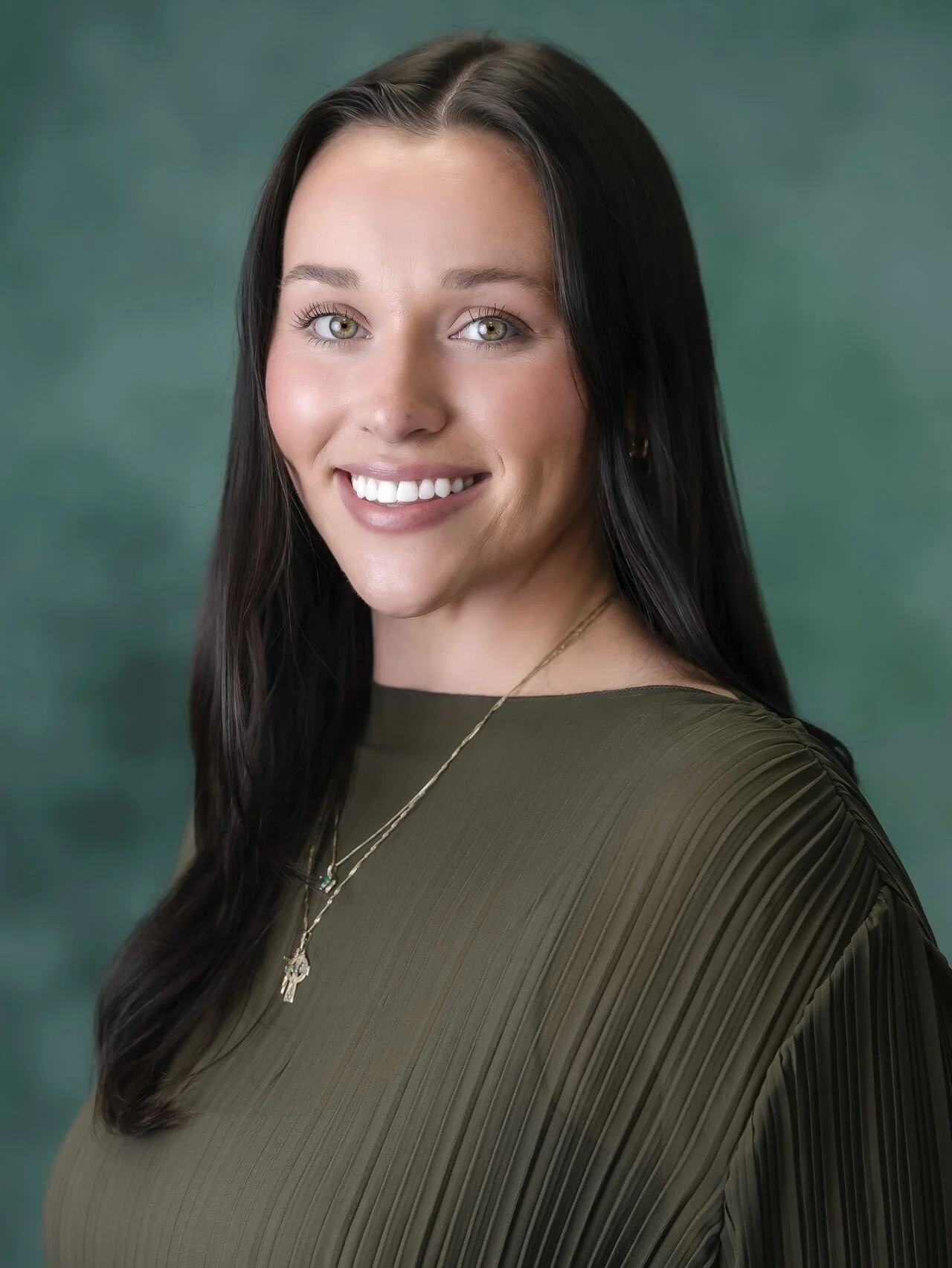 A woman with long dark hair, smiling, wearing a green top and a necklace with a cross pendant, in front of a green background.