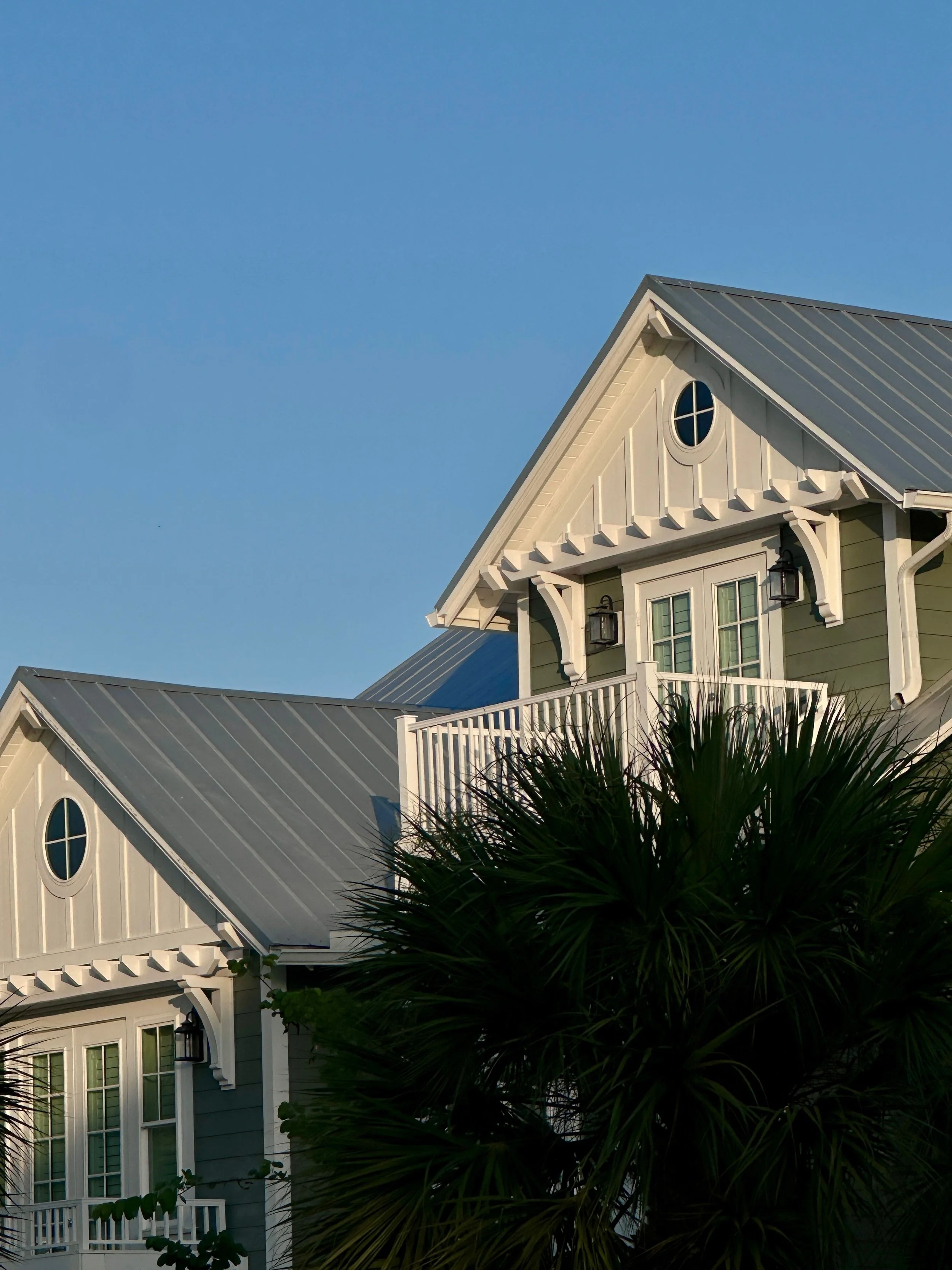 Close-up of a house with a gabled roof, white trim, and a small circular window, partially obscured by a large, dark green palm tree in front of it, under a clear blue sky.