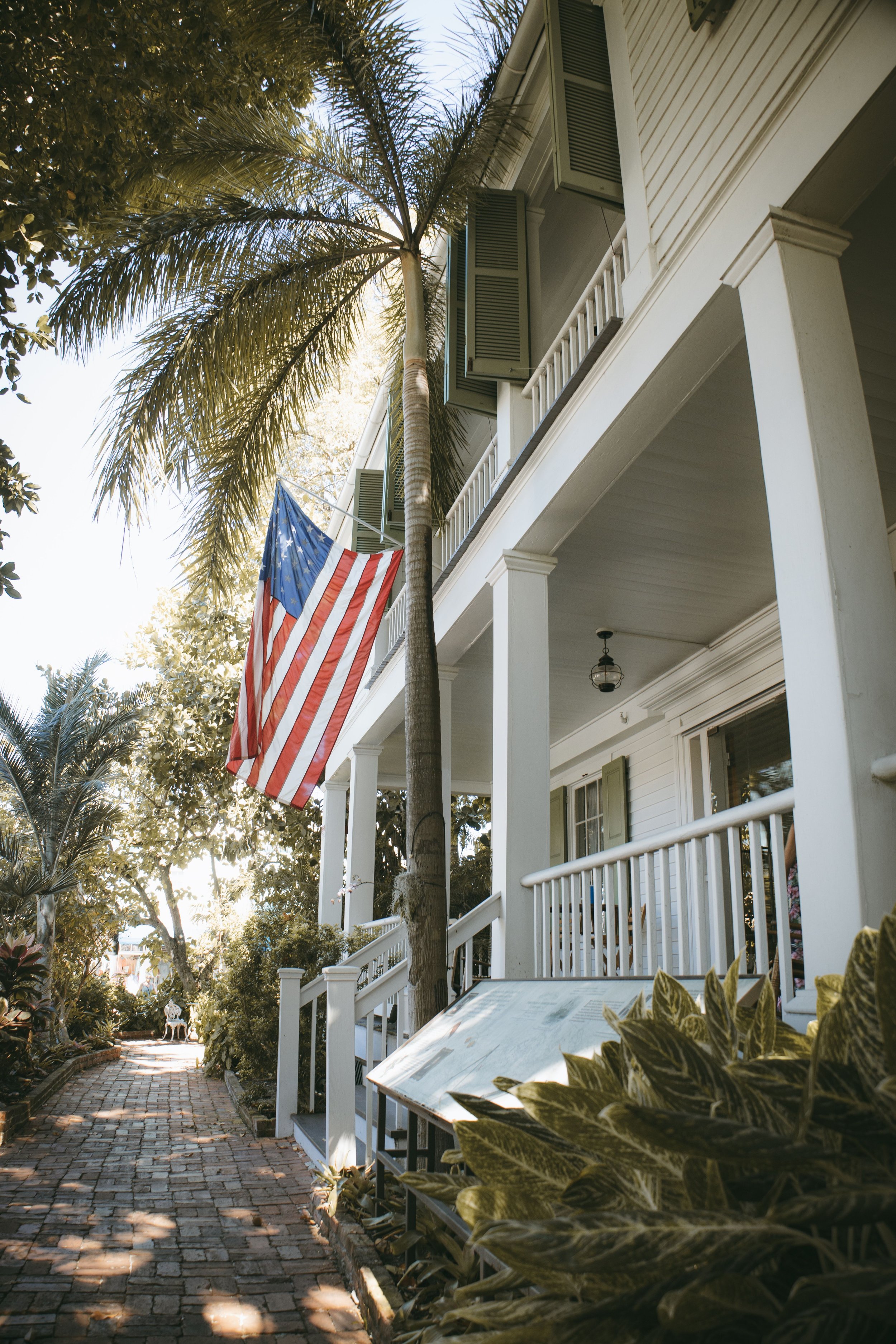 A white house with a porch and green shutters, a hanging lantern, and American flag, with a tall palm tree and lush greenery. A cobblestone pathway leads past a bench in the garden.