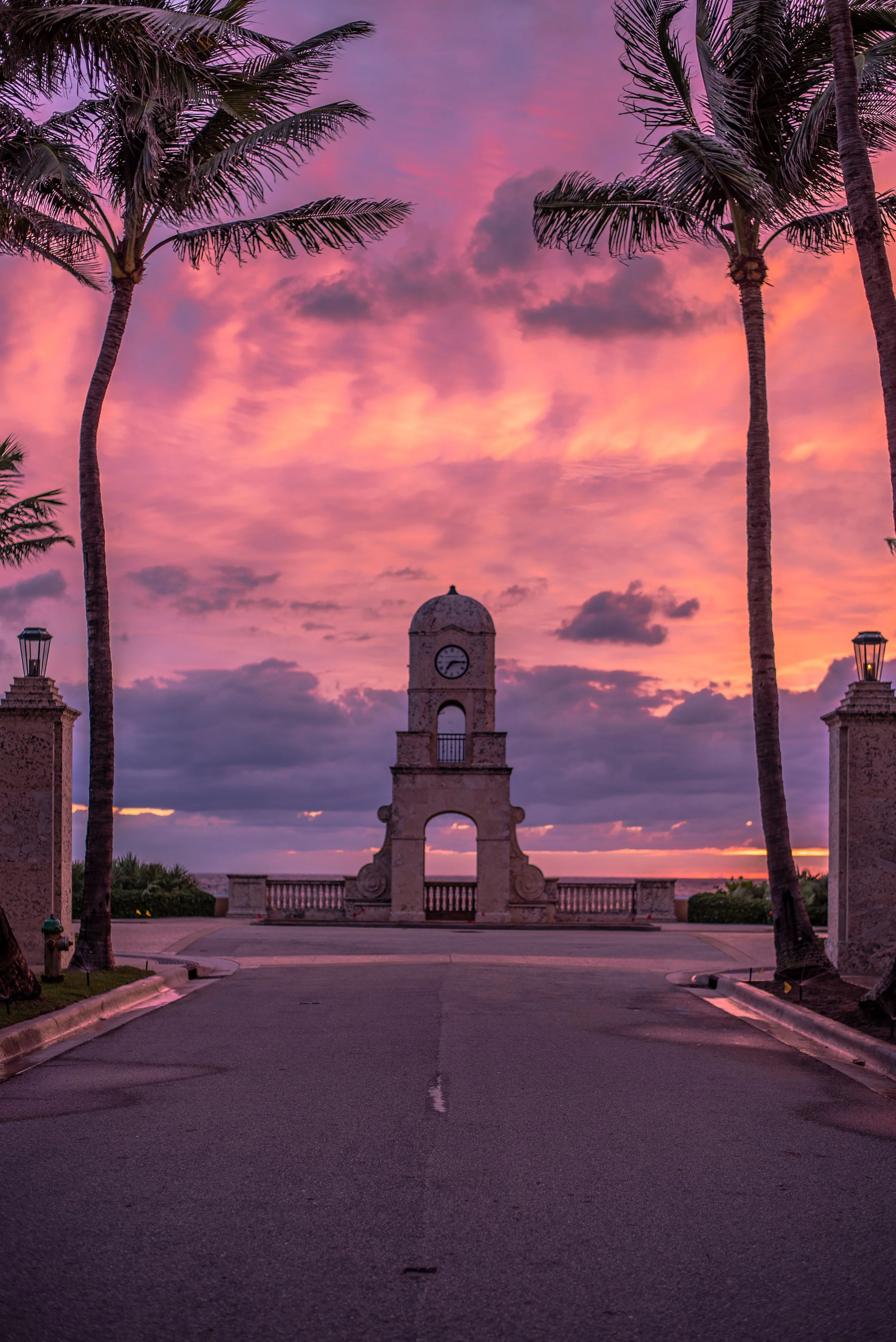 Sunset over a historic stone monument with a clock tower, flanked by two tall palm trees, in a coastal location.
