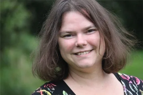 Portret van een vrouw met bruin haar, glimlachend, buiten met groene achtergrond.
