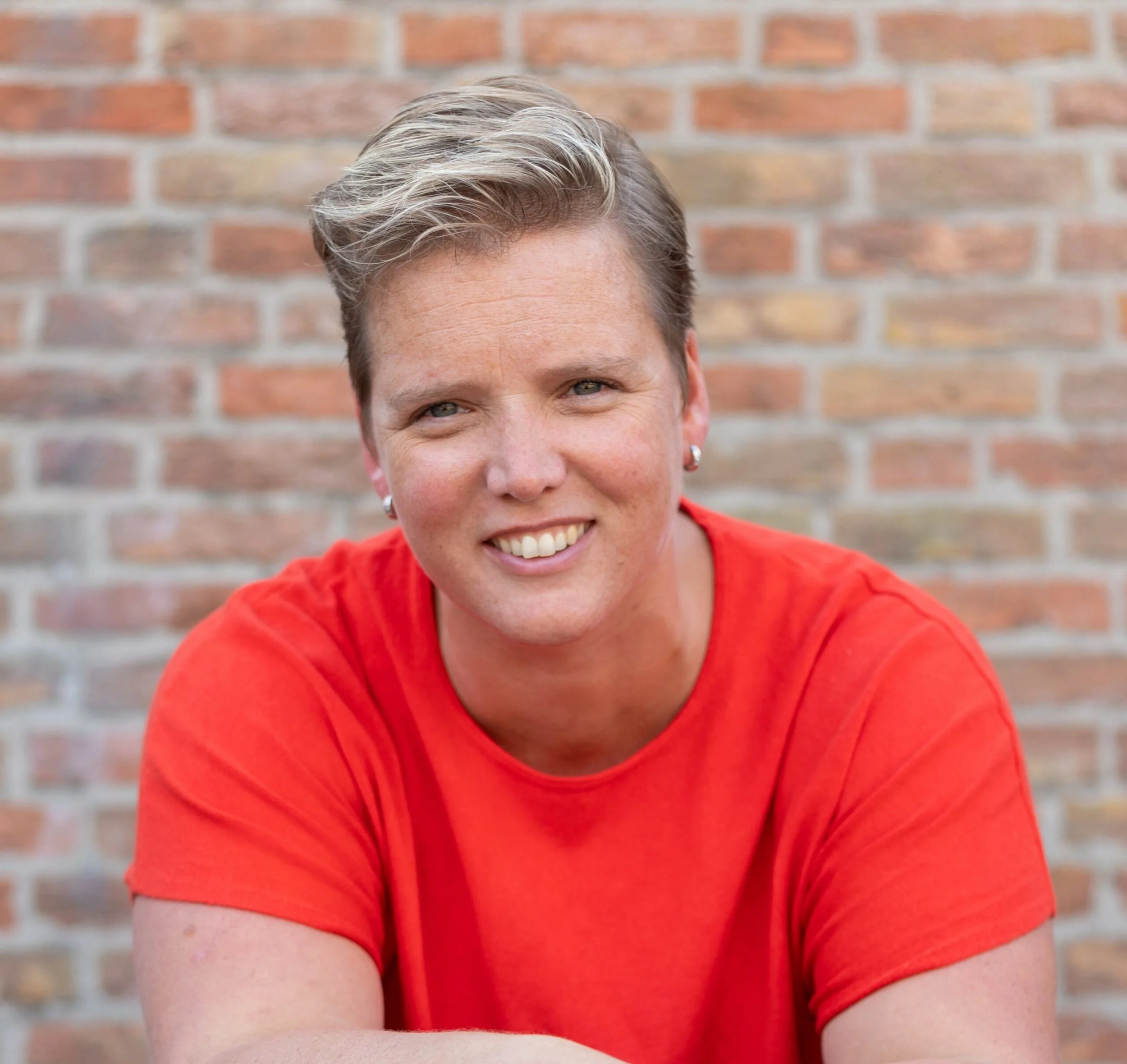 Close-up van een lachende vrouw met kort, zandkleurig haar, die een rode top draagt, voor een bakstenen muur.