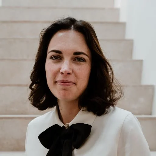 Portret van een jonge vrouw met bruin haar, witte blouse en een zwarte strik, staand op een trap in een witte omgeving.
