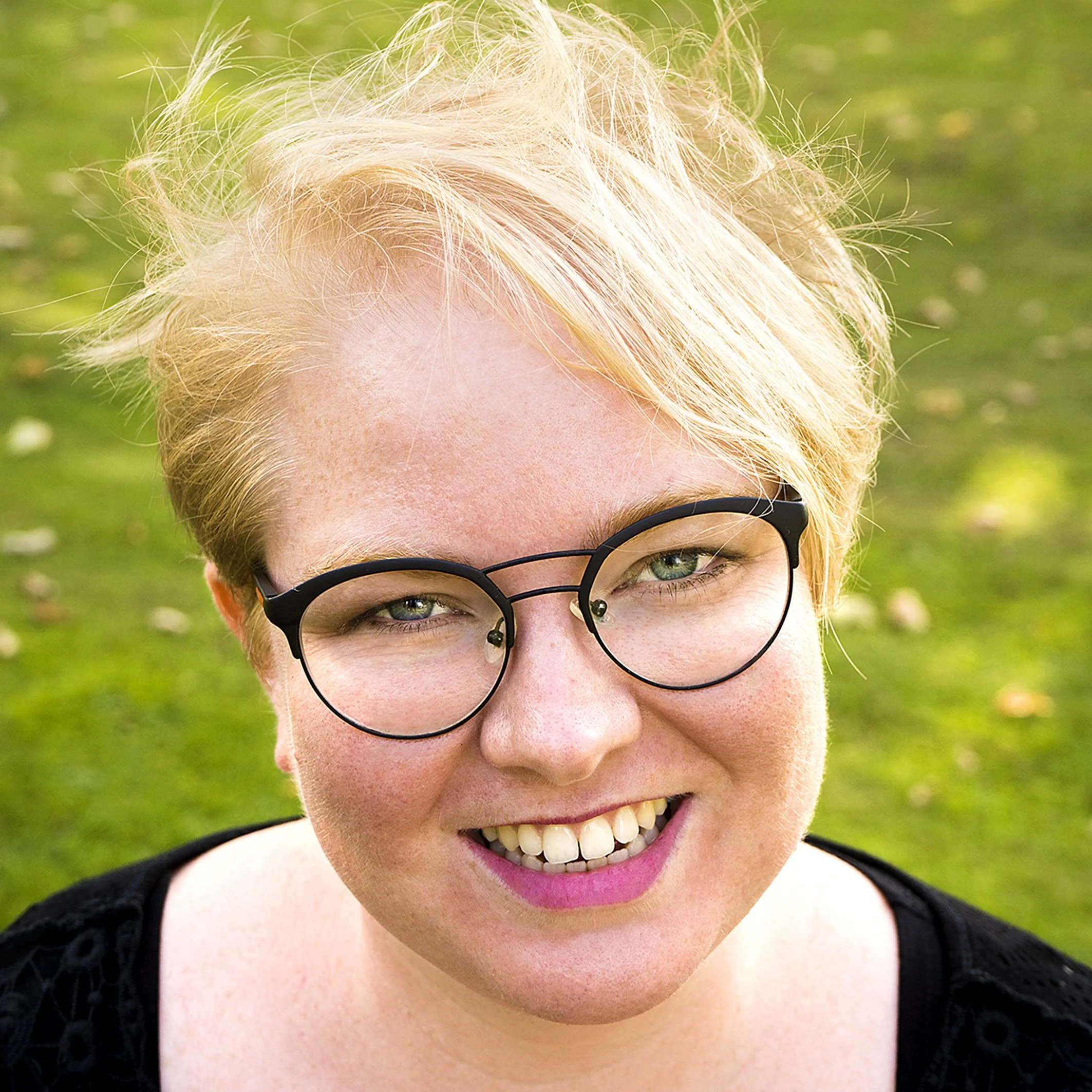 Close-up van een lachende vrouw met blond haar, zwarte bril en een glimlach, buiten met groene achtergrond.