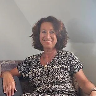 Vrouw met kort, krullend haar zit op een bank en lacht, draag een zwart-wit gekleed shirt.