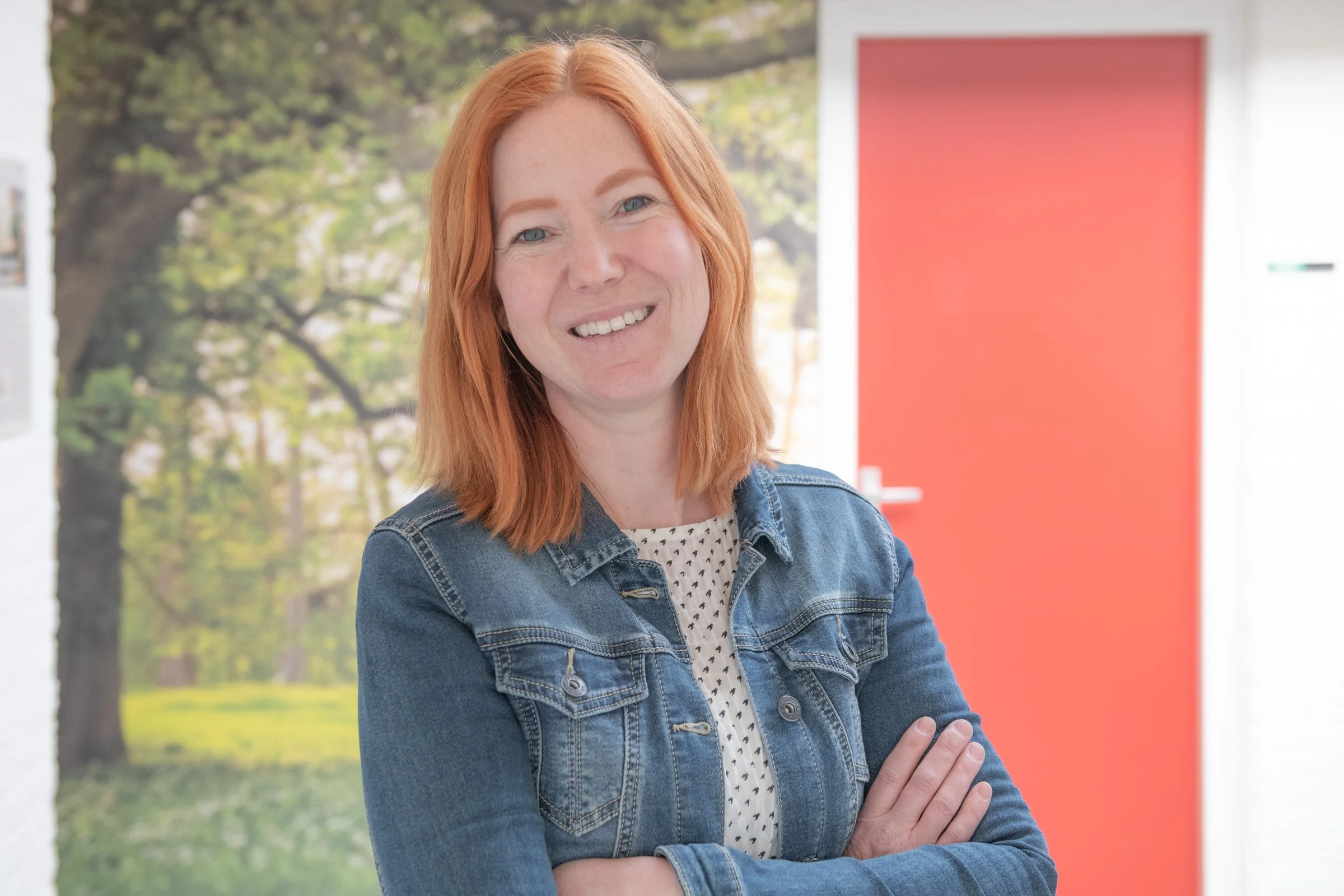 Vrouw met rood haar gekleed in een spijkerjack en een witte blouse met zwarte stippen, lachend en met gekruiste armen, binnen voor een muur met een achtergrond van een bos en een rood paneel.