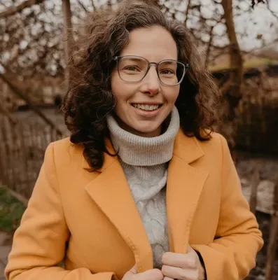 Vrouw met bril, krullend haar, gele jas en grijze trui, lachend buiten in een tuin met herfstbladeren.