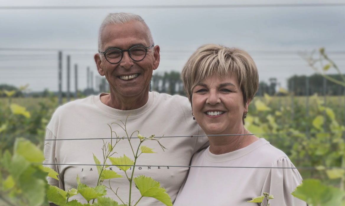 Een oudere man en vrouw glimlachen in een wijngaard, omringd door groene wijnrankplanten.