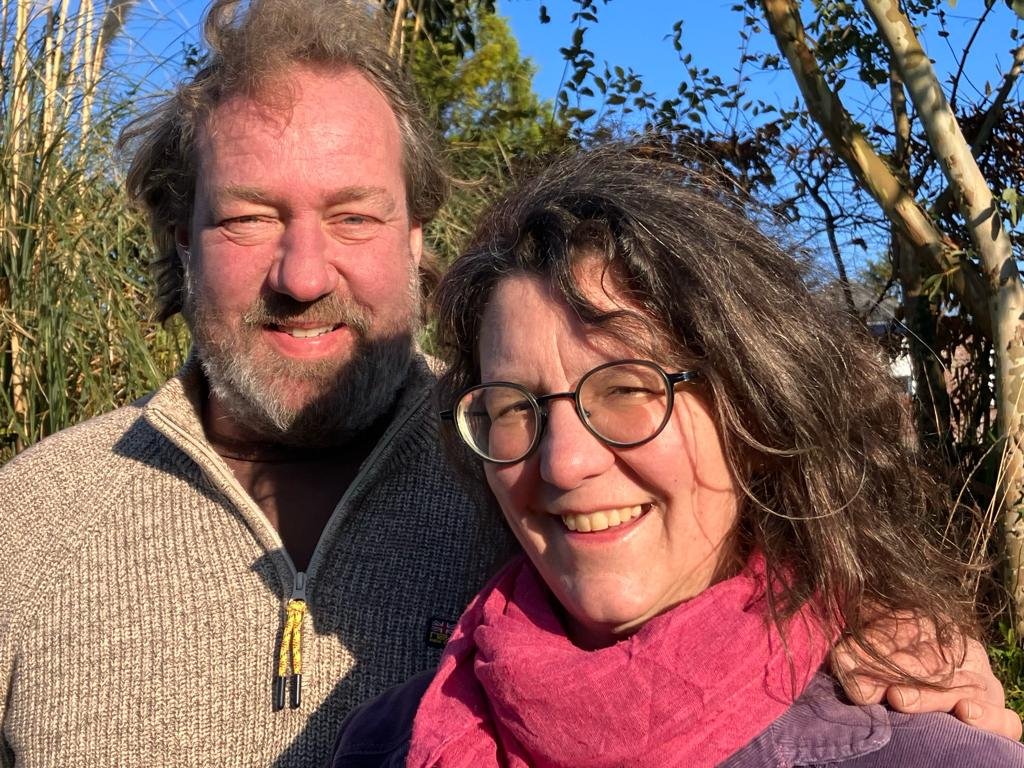 Twee mensen, een man met een bebaarde, grijze haar en een vrouw met golvend haar en brillen, staan buiten in de zon, omringd door planten en bomen, en lachen.