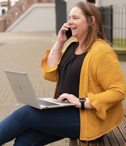 Inge van Wepzz die buiten op een bank zit, met een laptop op haar schoot en een telefoon tegen haar oor, lachend en pratend. Inge helpt ondernemers met online marketing vraagstukken.