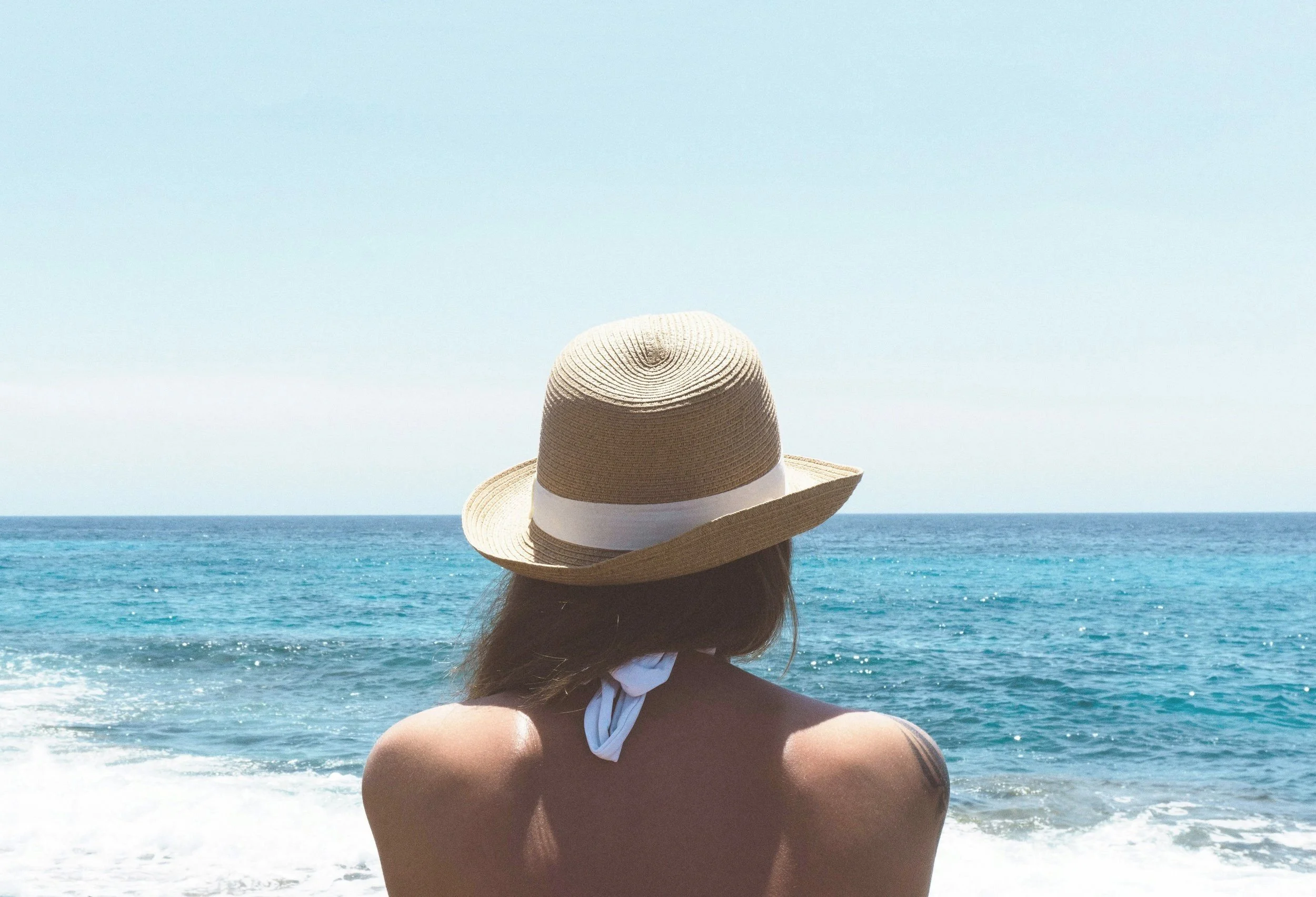 A person wearing a straw hat facing the ocean.