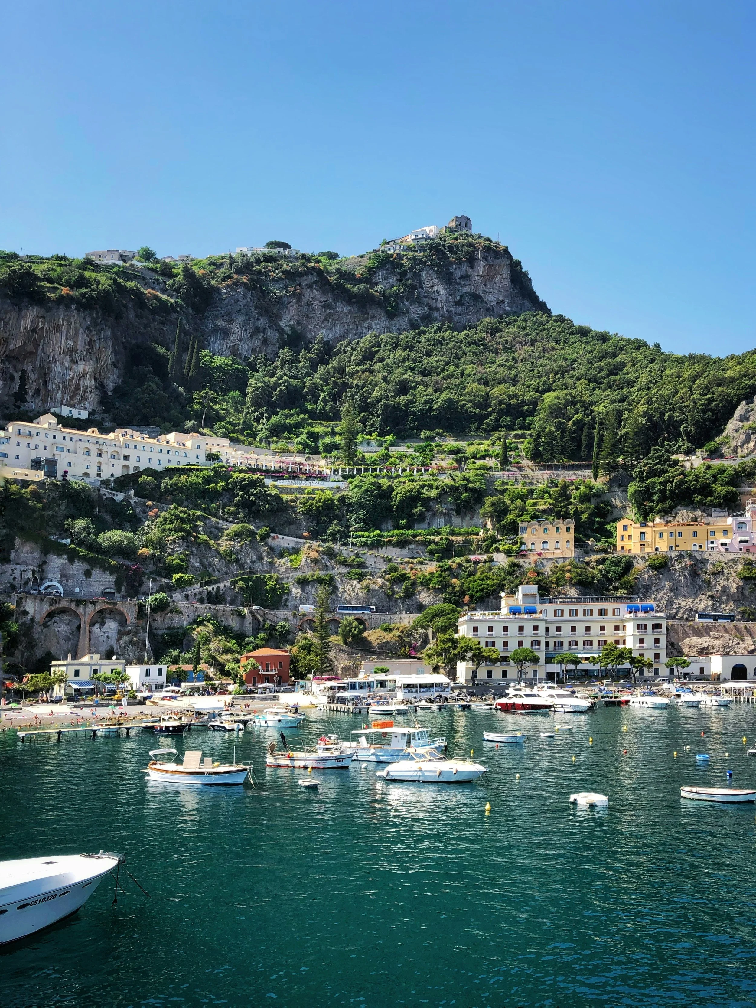 Coastal scene with boats on the water, buildings, and lush green mountains, with a clear blue sky.