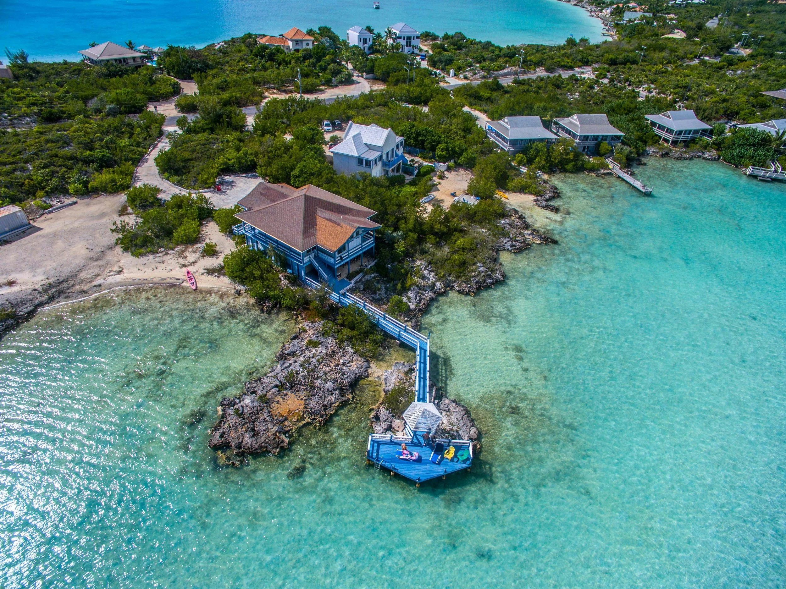Aerial view of a coastal area with several houses, surrounded by clear turquoise waters and green vegetation. A wooden deck extends over the water, with a small gazebo and chairs. The landscape includes rocky shores and sandy patches.