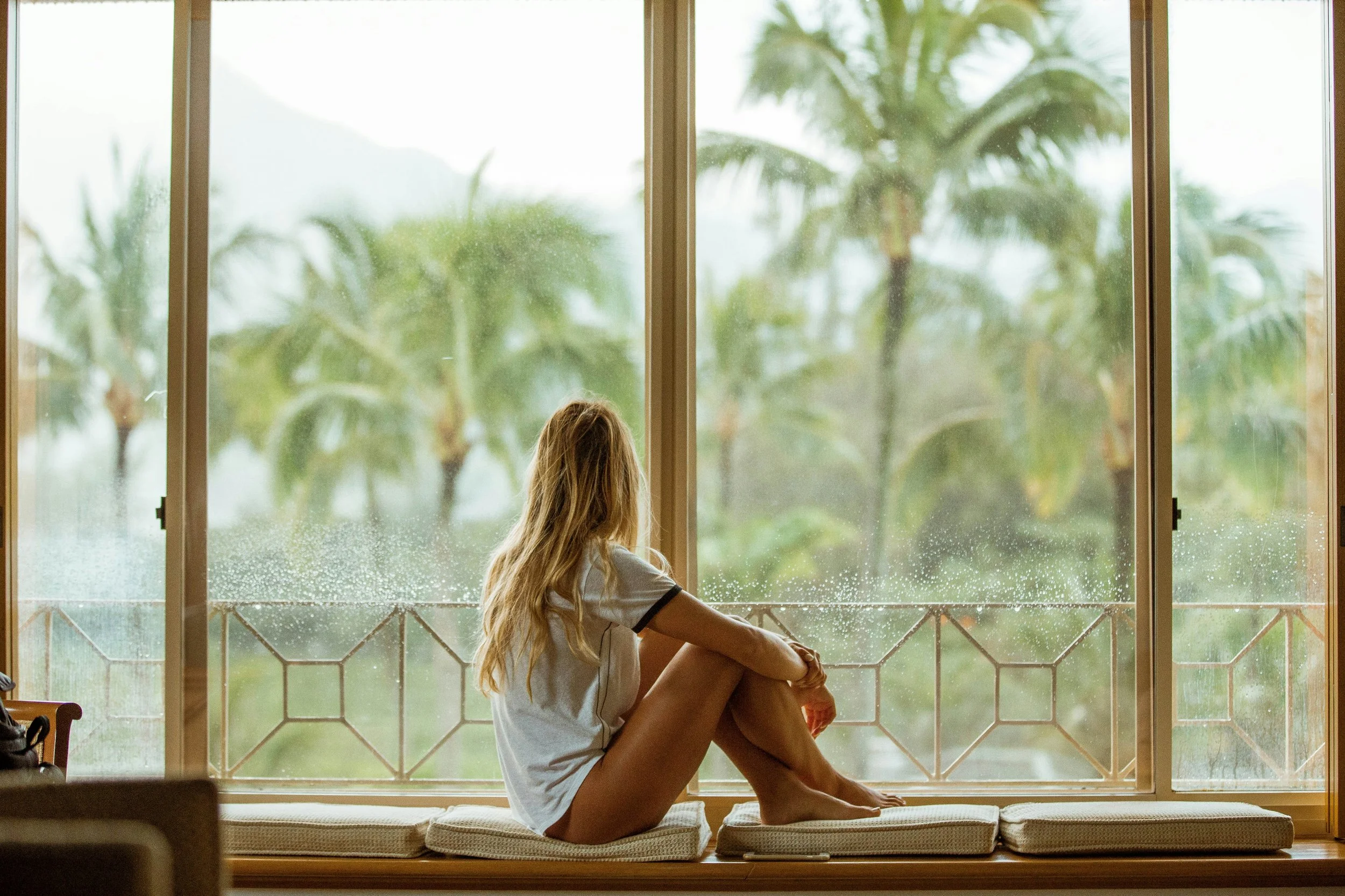 A young woman with long blonde hair sitting on a windowsill and looking outside on a rainy day. The window shows rain and palm trees outside.