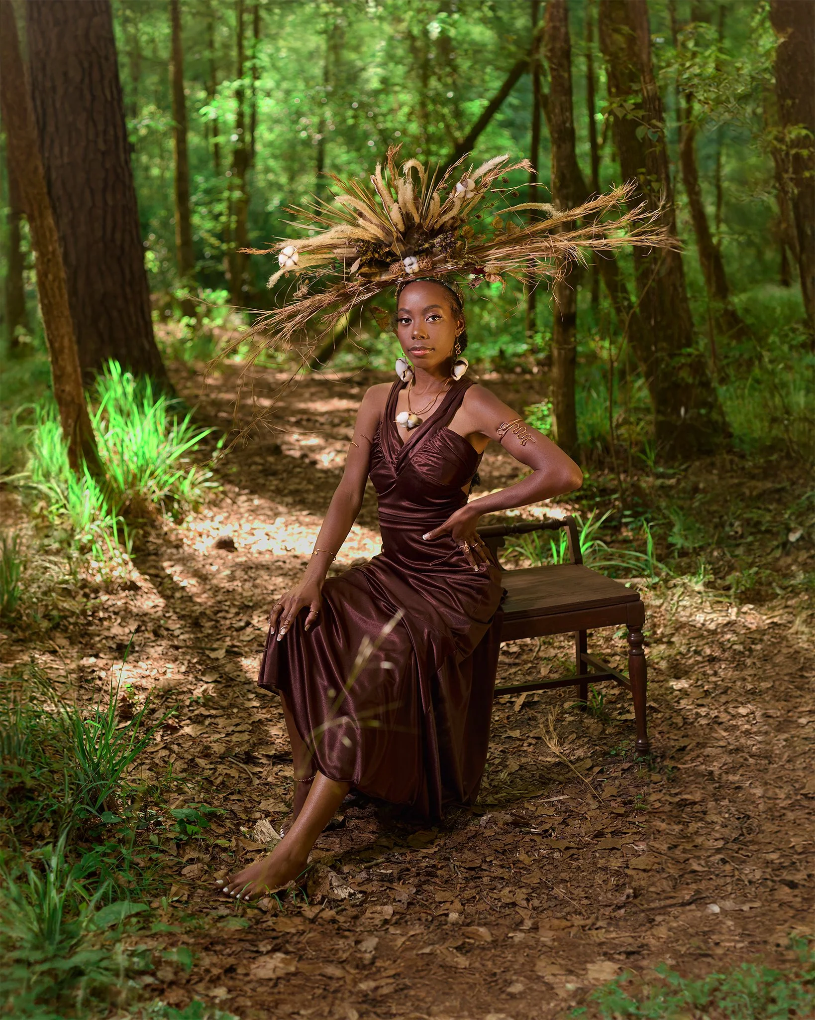 A woman sits on a wooden bench in a forest, wearing an elegant brown satin dress and an elaborate headpiece decorated with dried plants, shells, and natural elements. She has a confident expression, adorned with jewelry, and is posing with one arm resting on her waist.
