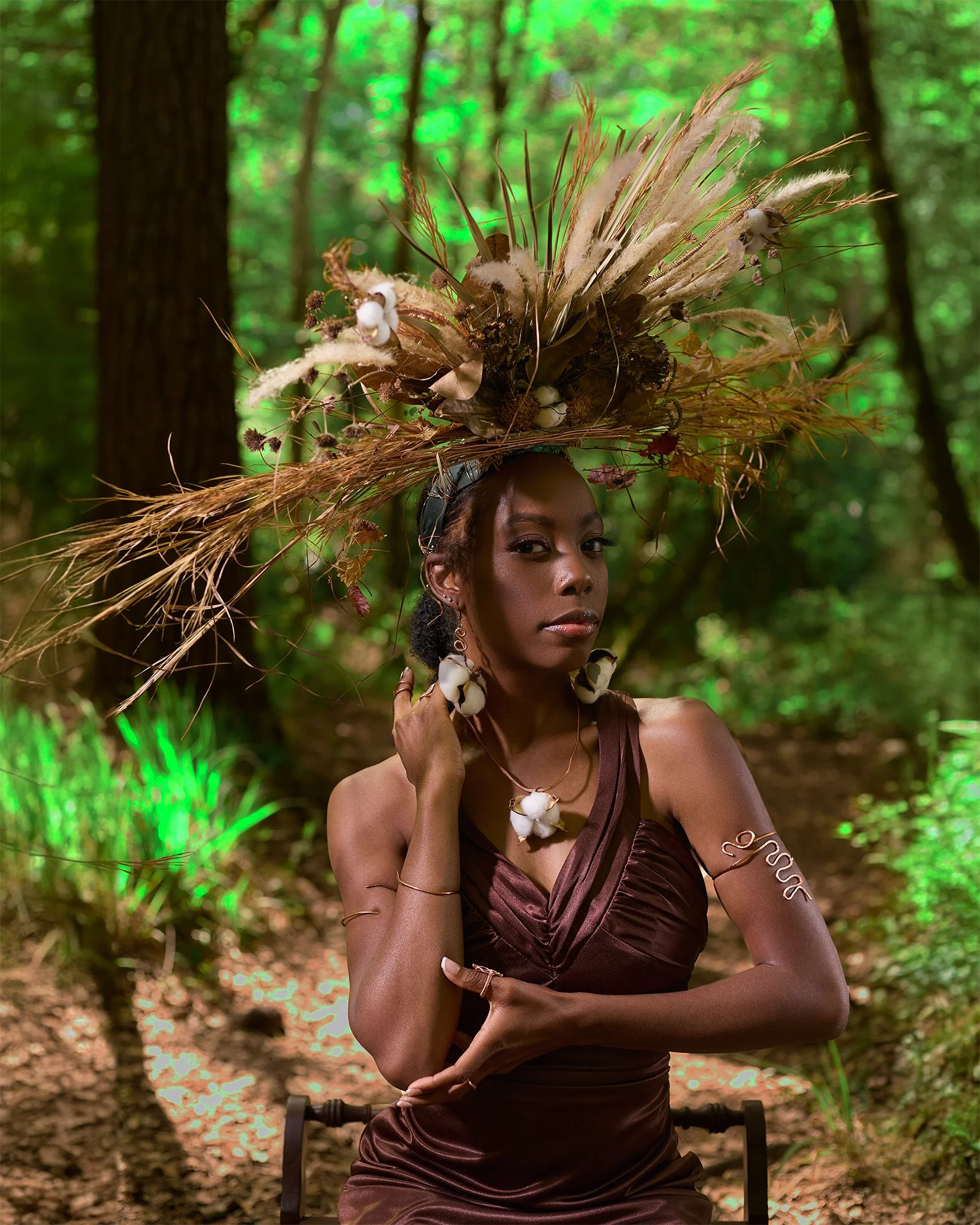 A woman in a brown satin dress with ornate jewelry, seated outdoors in a forest, wearing an elaborate headpiece made of dried plants, feathers, and cotton-like flowers.