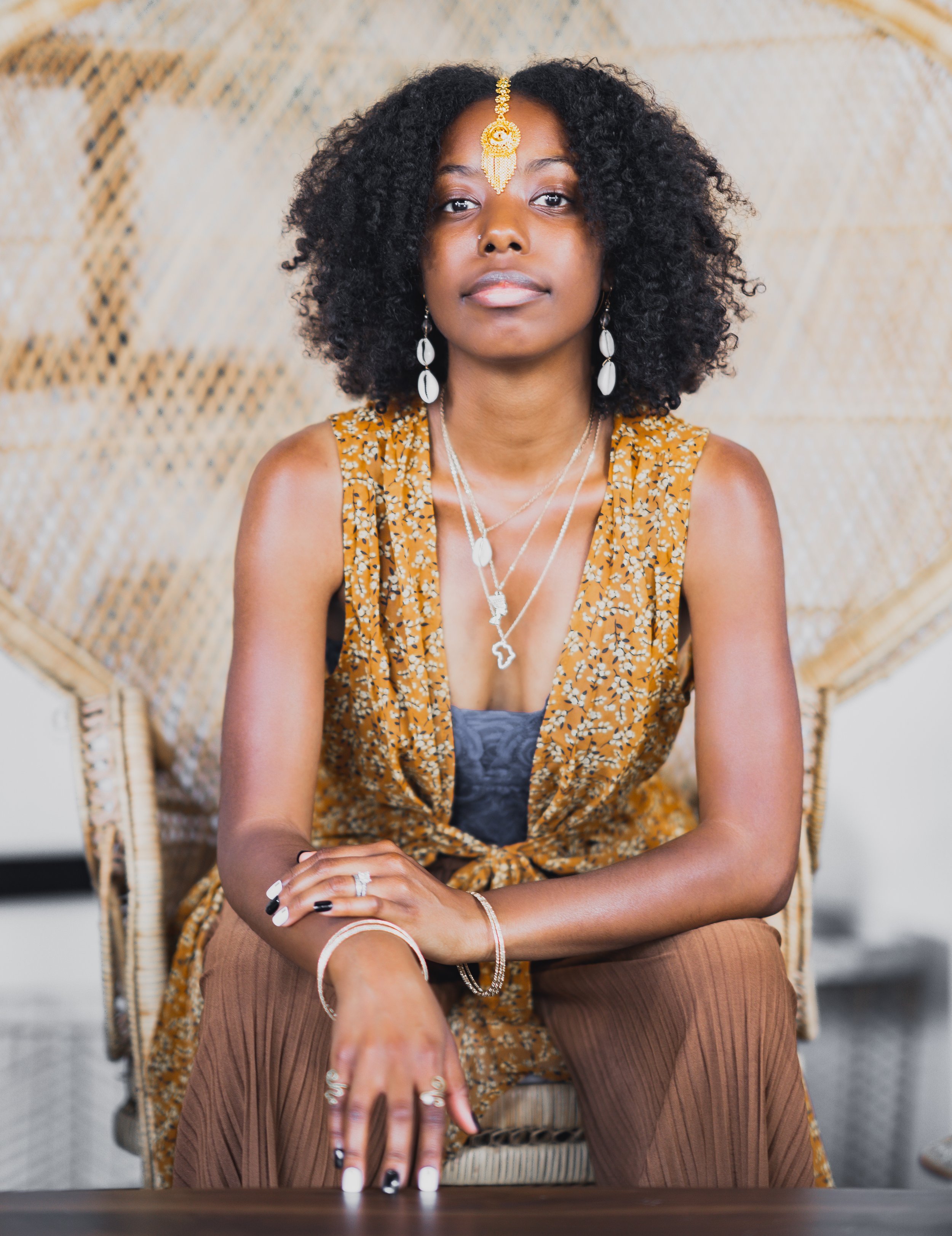 A young woman with curly black hair, wearing jewelry including earrings, necklaces, and a gold forehead chain, sitting in front of an ornate golden, woven background.