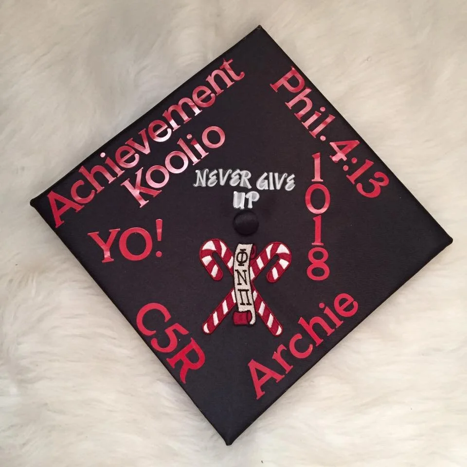 Graduation cap decorated with red and white theme, displaying phrases like "Achievement," "Never Give Up," "YO!" and the name "Archie." It features a candy cane design and pandemic-related message "Phil 4:13" along with the graduation year 2018.