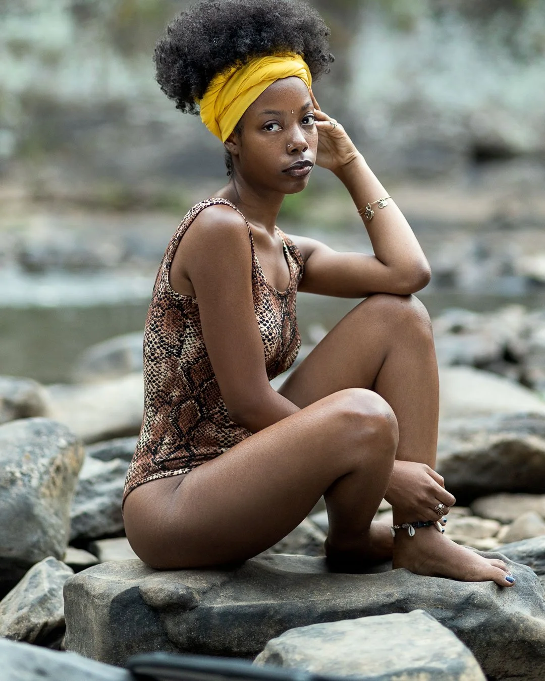 A woman with dark skin and natural curly hair sitting on rocks by a river, wearing a leopard print sleeveless top, a yellow headscarf, and jewelry.