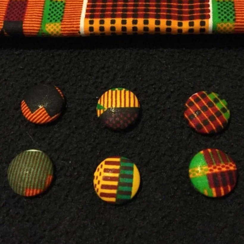 Six round fabric-covered buttons with colorful plaid and striped patterns, placed on a black surface beneath a fabric with a similar pattern.