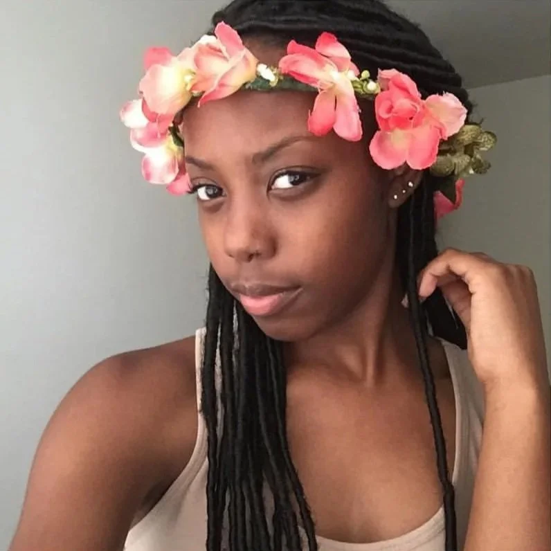 A young woman with dark skin and long dreadlocks wearing a floral headband with pink and peach flowers, posing with her hand near her face against a plain background.