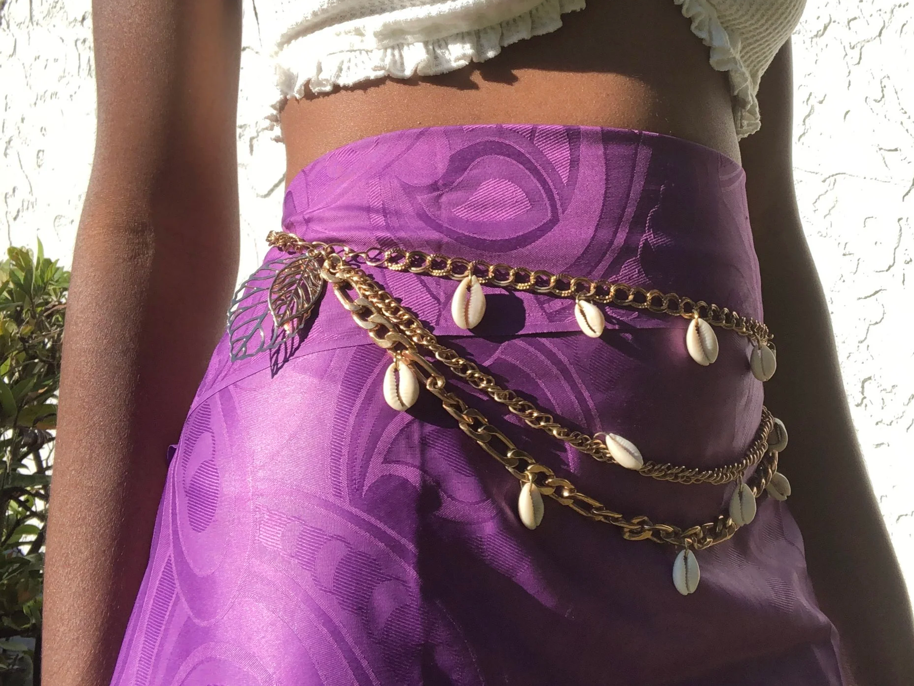 Close-up of a woman’s midsection showcasing a purple skirt with a subtle floral pattern, adorned with layered gold chains and shell charms, against a white textured wall background.