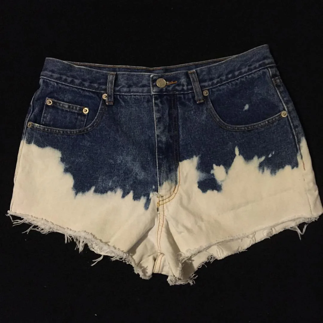 A pair of denim shorts with a distressed, frayed hem, featuring a faded, bleached pattern on the lower part of the legs against a black background.
