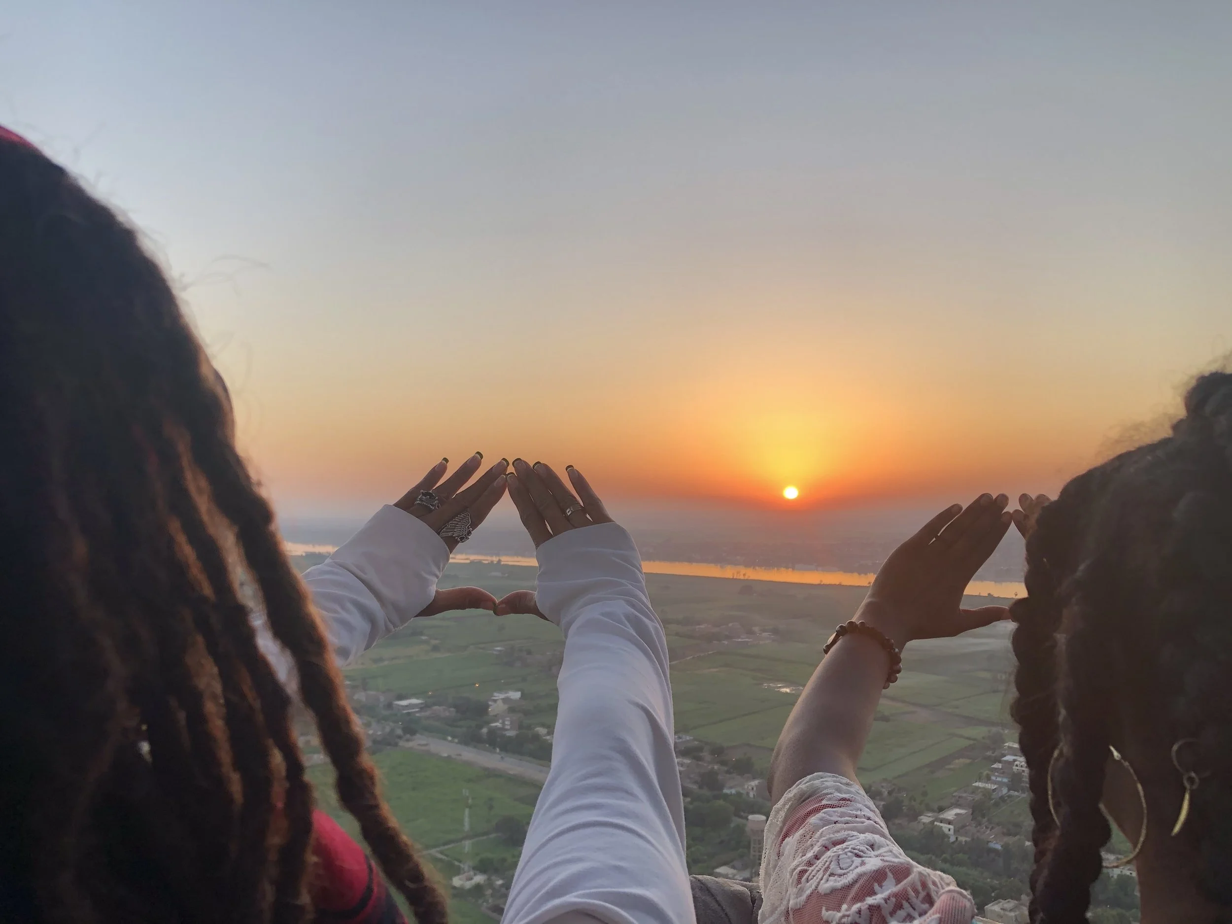 People celebrating sunset outdoors, making a heart shape with hands, overlooking a landscape with fields, a river, and buildings.