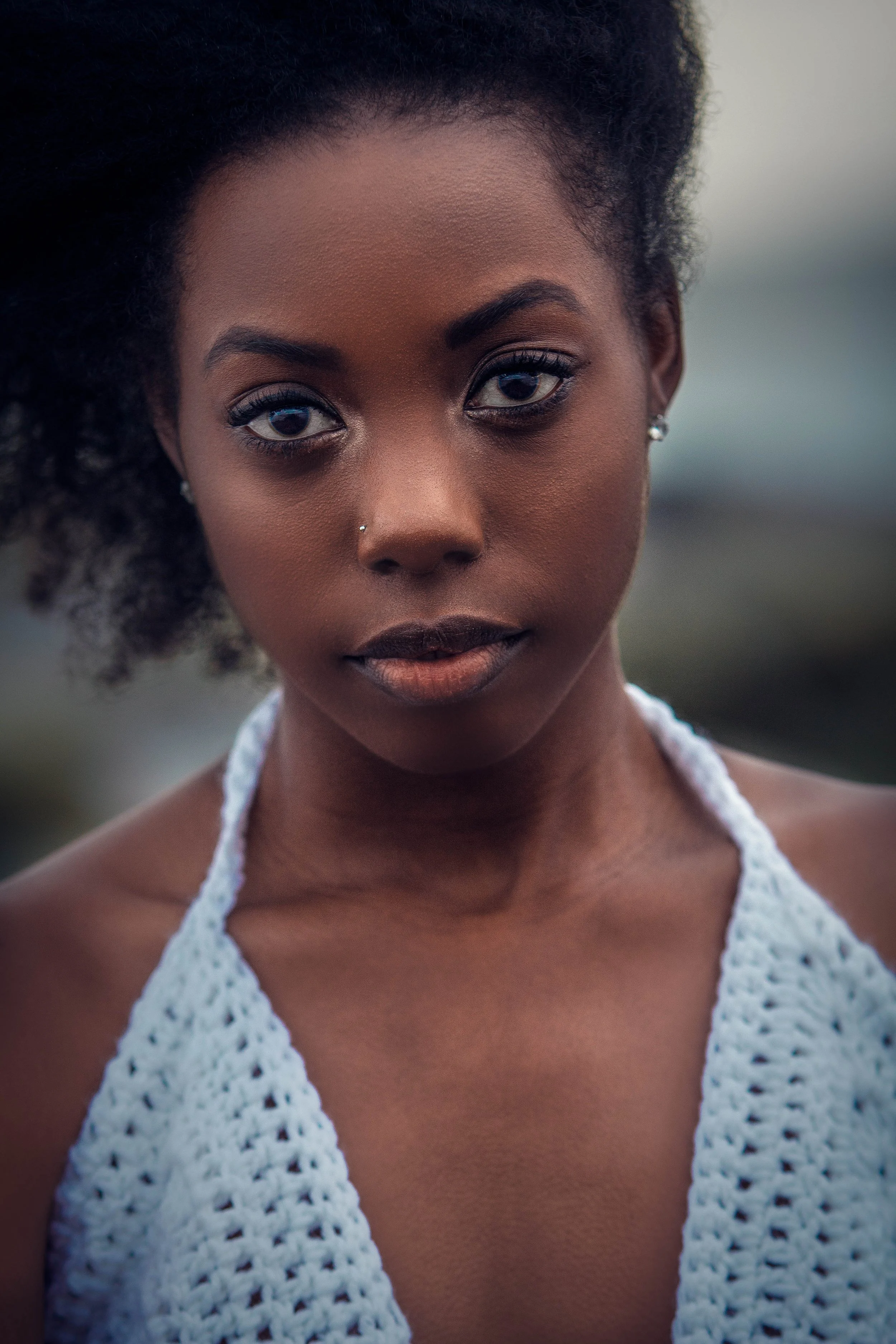 Close-up portrait of a young woman with dark skin, curly black hair, and a nose piercing, wearing a white crocheted top.
